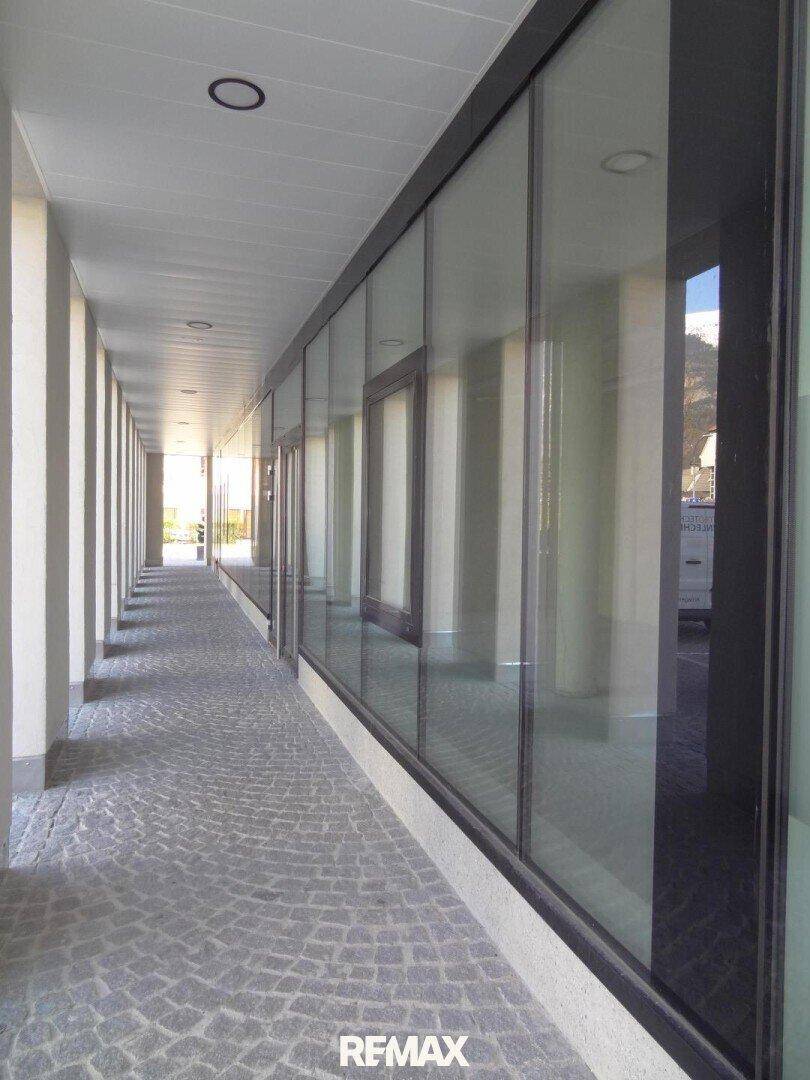 Langer, überdachter Gang entlang einer modernen Glasfassade mit gepflastertem Boden und Säulen.