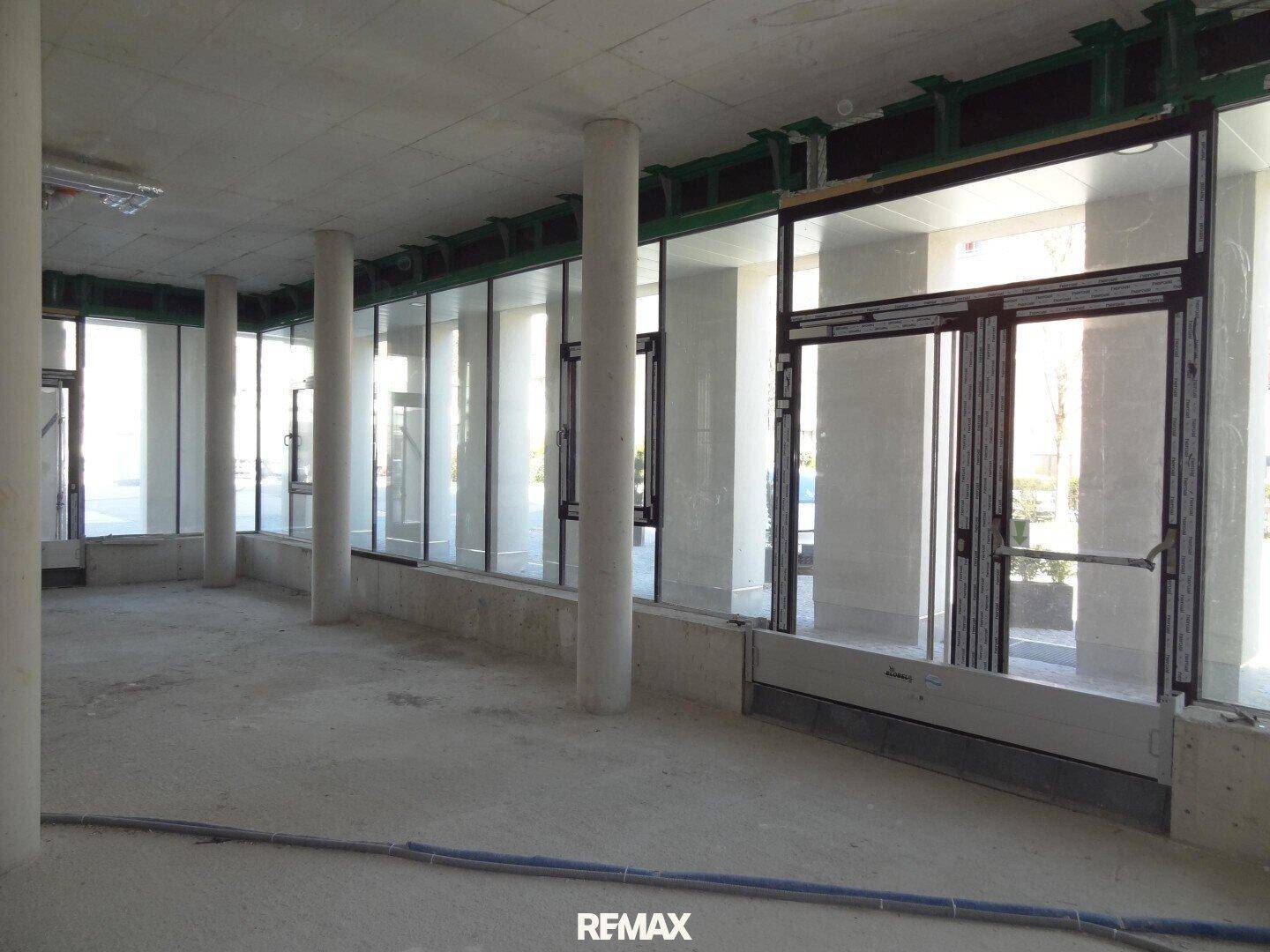 Unfertiger Innenraum eines Geschäftsraums mit hohen Decken, Betonboden und großen Fensterfronten für viel Tageslicht.
