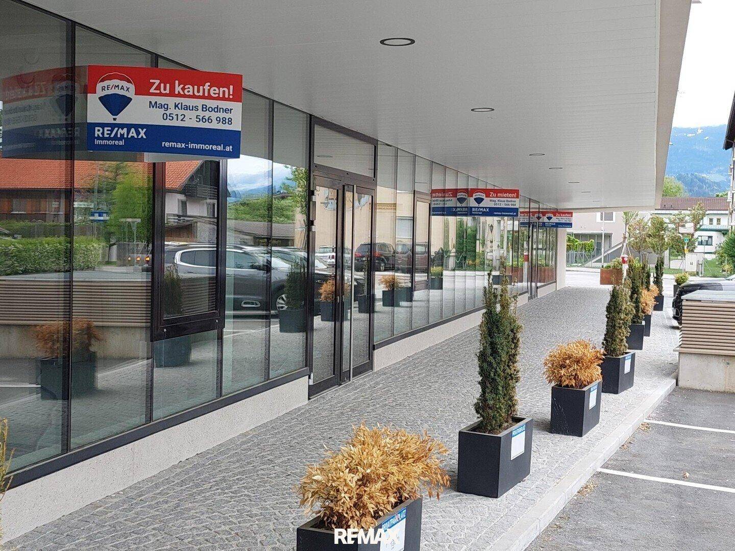 Erdgeschoss mit großen Glasfronten und Werbeschildern, umgeben von gepflasterten Flächen und Pflanzen.