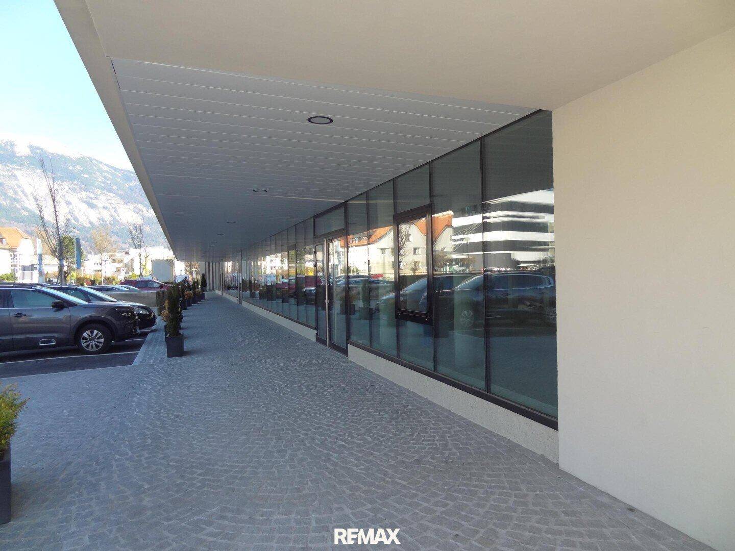 Moderne Gebäudefassade mit durchgehender Glasfront, gepflastertem Gehweg und Parkplätzen im Vordergrund.