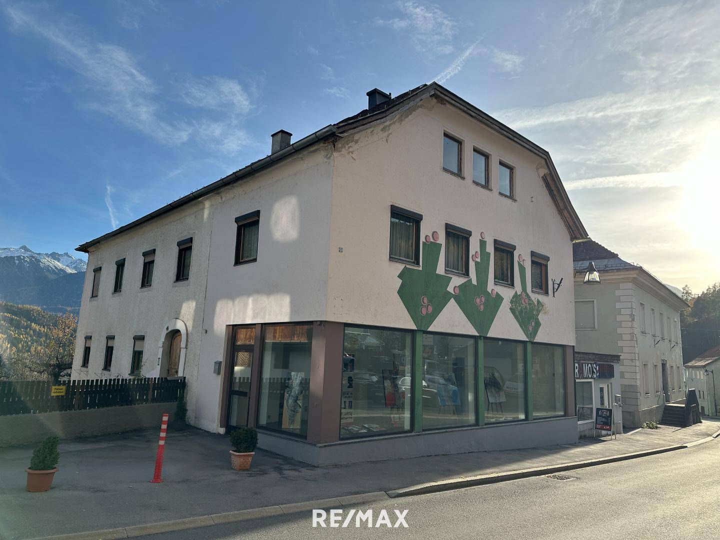 Gebäude mit Ladenfront im Erdgeschoss und Wohnungen darüber, umgeben von Bergen unter blauem Himmel.