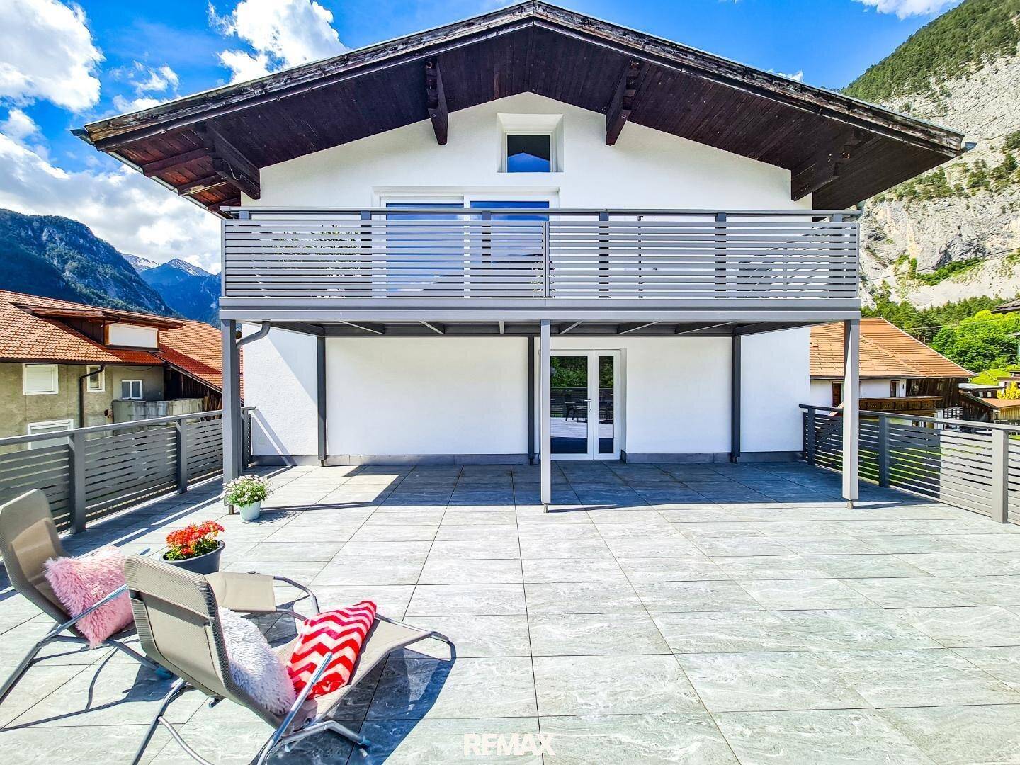 Hausfassade mit großer Terrasse im Erdgeschoss und Balkon im Obergeschoss, umgeben von Bergen.