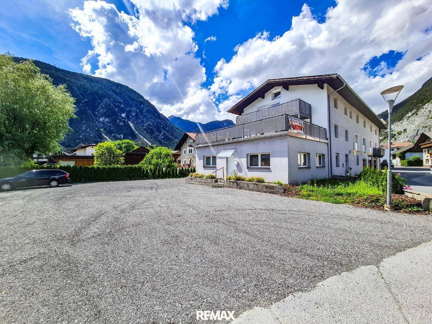 Mehrfamilienhaus mit großem Schotterparkplatz und Bergblick unter blauem Himmel.