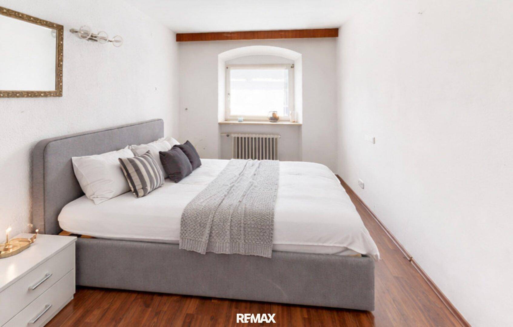 Schlafzimmer mit grauem Polsterbett, Holzboden und Fenster für angenehmes Wohnen.