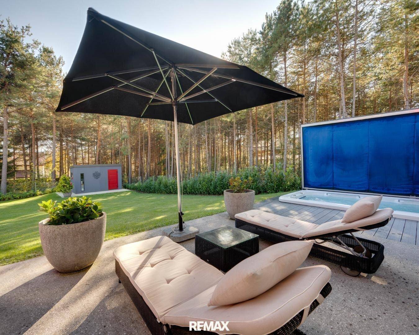 Großzügige Terrasse mit Whirlpool, Liegestühlen und umgeben von hohen Bäumen und Rasenfläche.