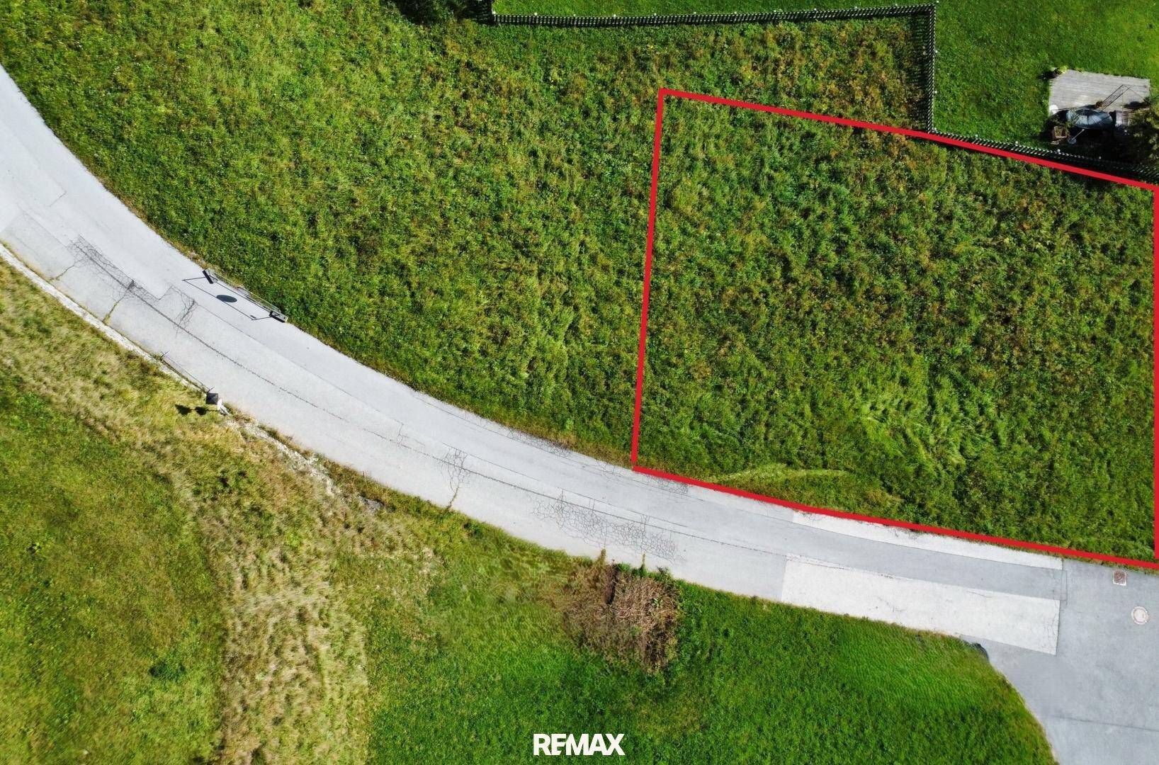 Luftaufnahme eines grünen Grundstücks, rot umrandet, neben einer Zufahrtsstraße in ländlicher Umgebung.