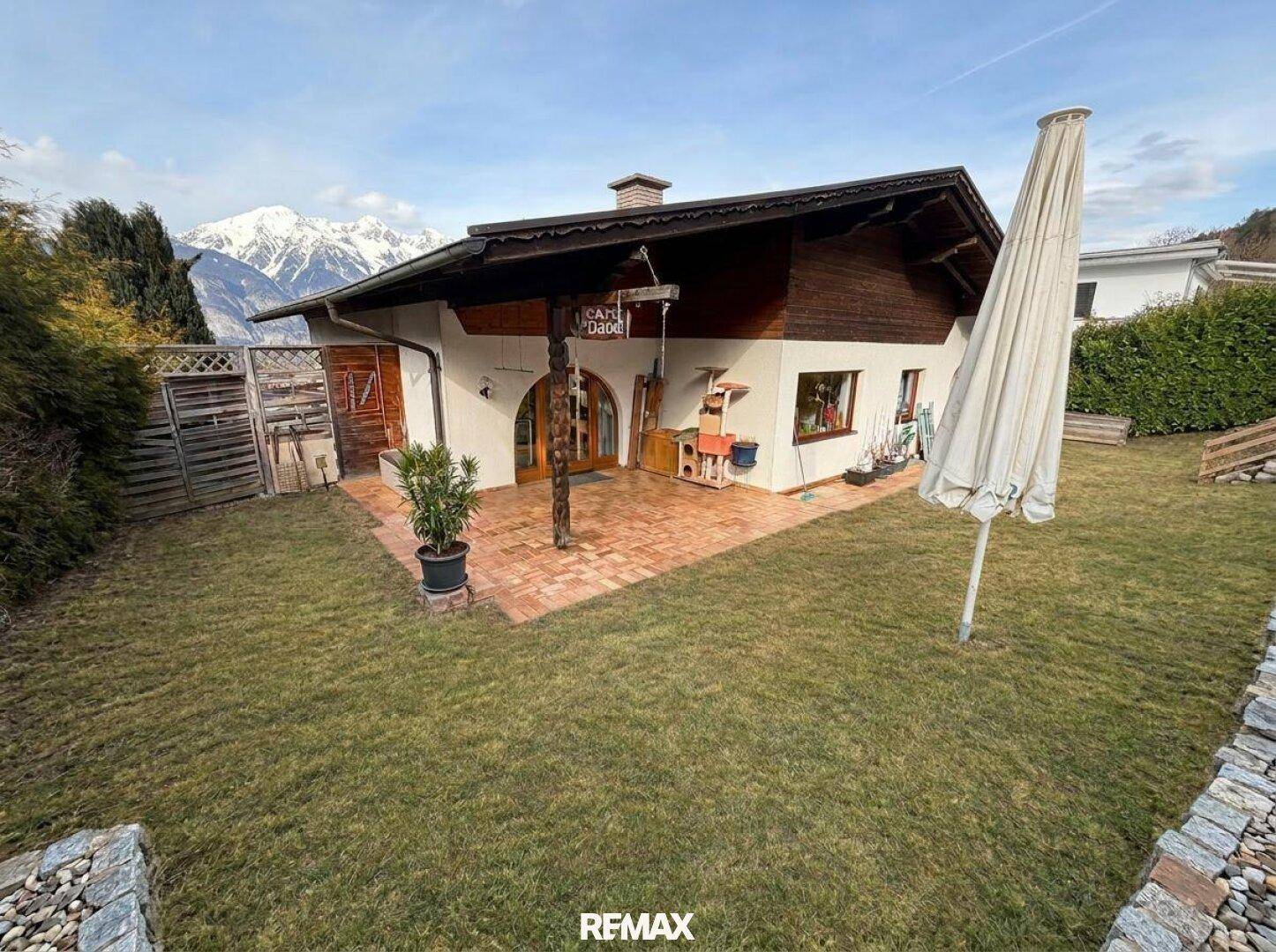 Gemütliche Terrasse am Haus mit Sonnenschirm und Topfpflanzen, umgeben von einem grünen Garten.