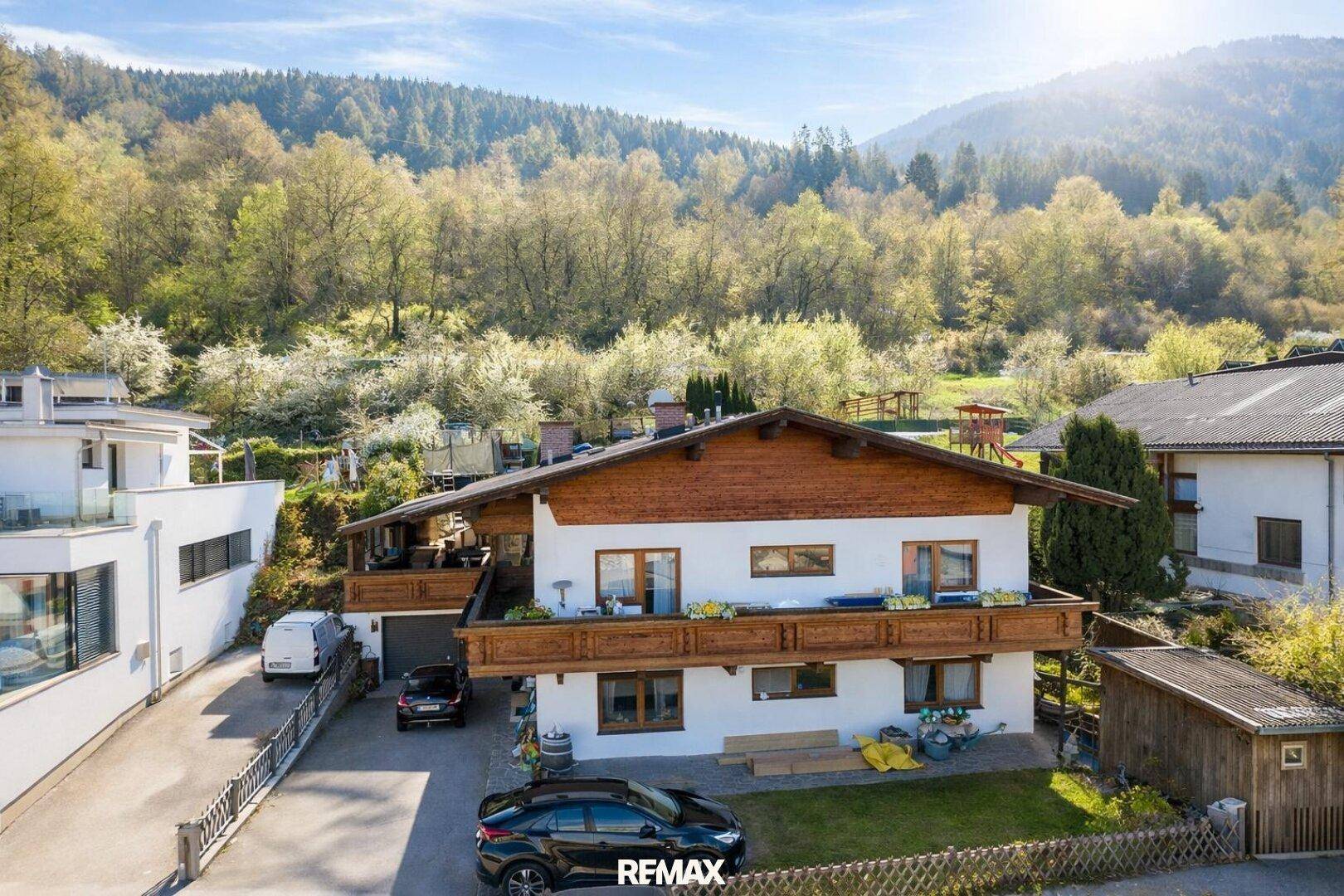Haus mit Holzbalkonen und gepflegtem Vorgarten, umgeben von grünen Hügeln und Bäumen.
