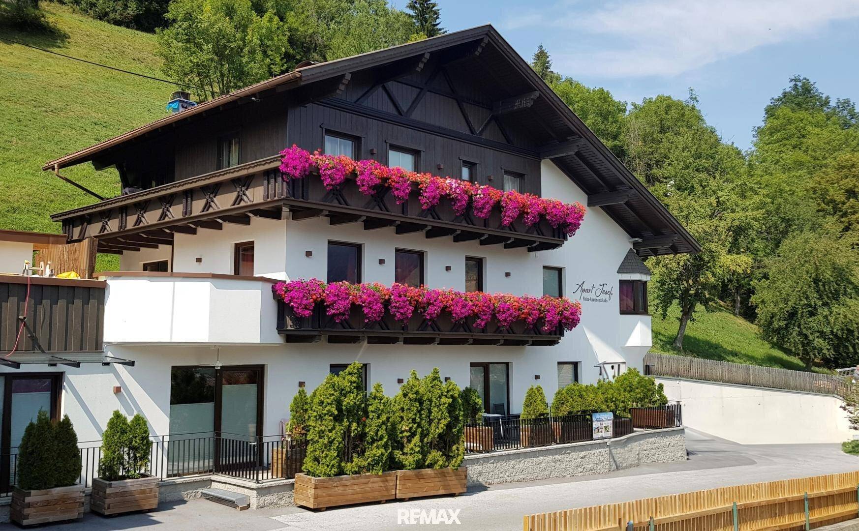 Traditionelles Gebäude mit weißer Fassade, dunklem Holzdach und blühenden Blumenkästen an den Balkonen.