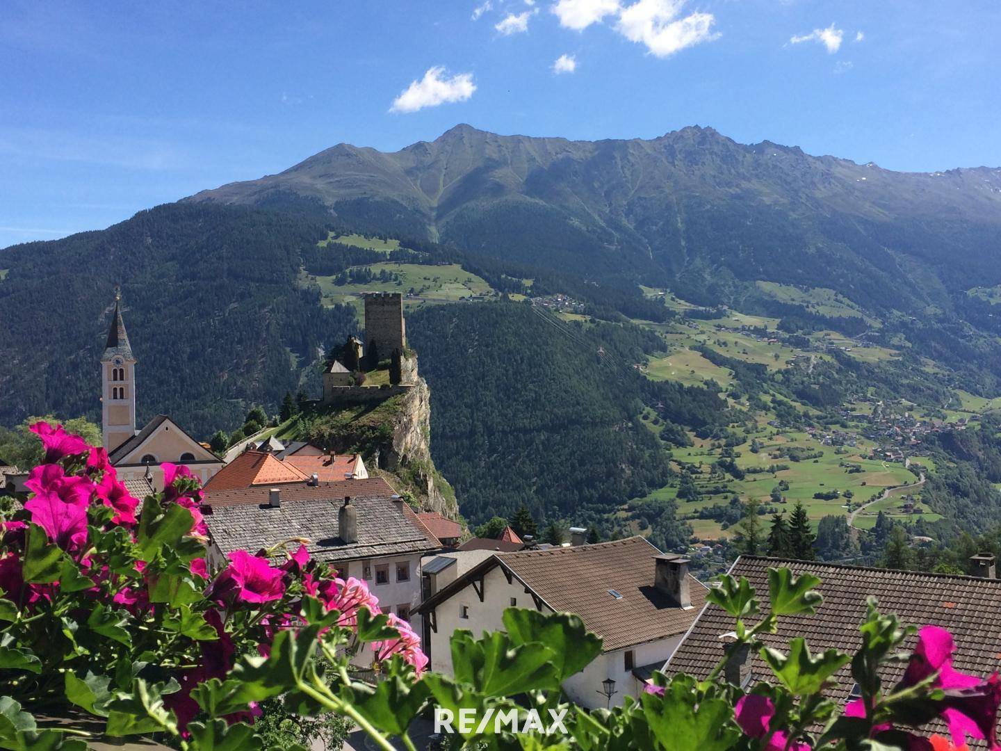 Malerischer Blick auf ein Bergdorf mit Burgruine und blühenden Blumen im Vordergrund.