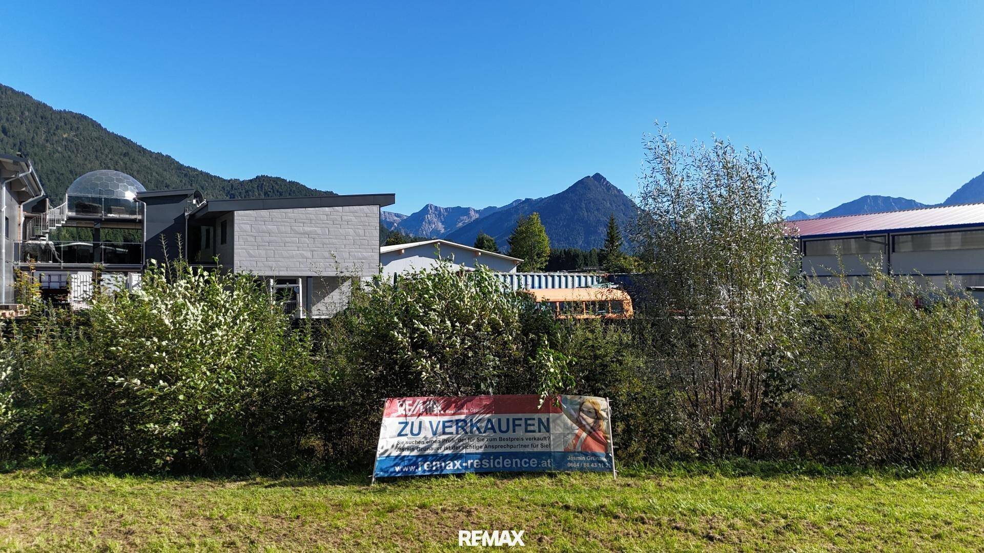 Ein "Zu Verkaufen"-Schild steht auf einer grünen Wiese vor einer Bergkulisse und Gebäuden.