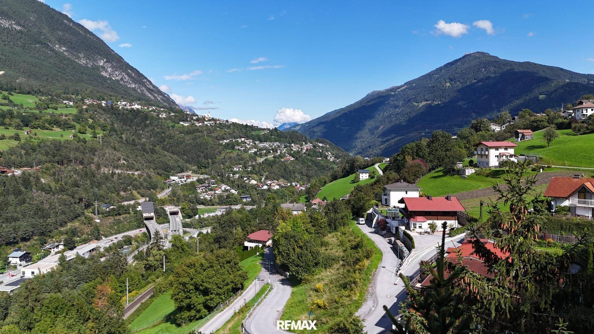 Weitläufiger Blick auf ein Tal mit einem Dorf, Straßen und imposanten Bergen unter einem klaren Himmel.
