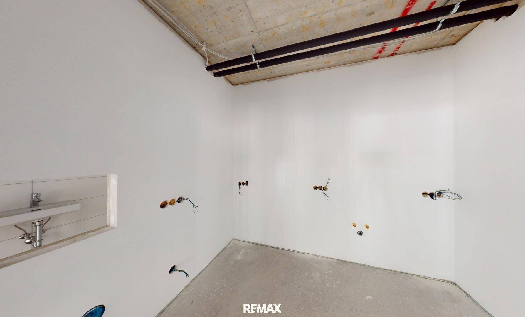 Badezimmer im Rohbau mit sichtbaren Installationen, Betonboden und einem kleinen Waschbecken.