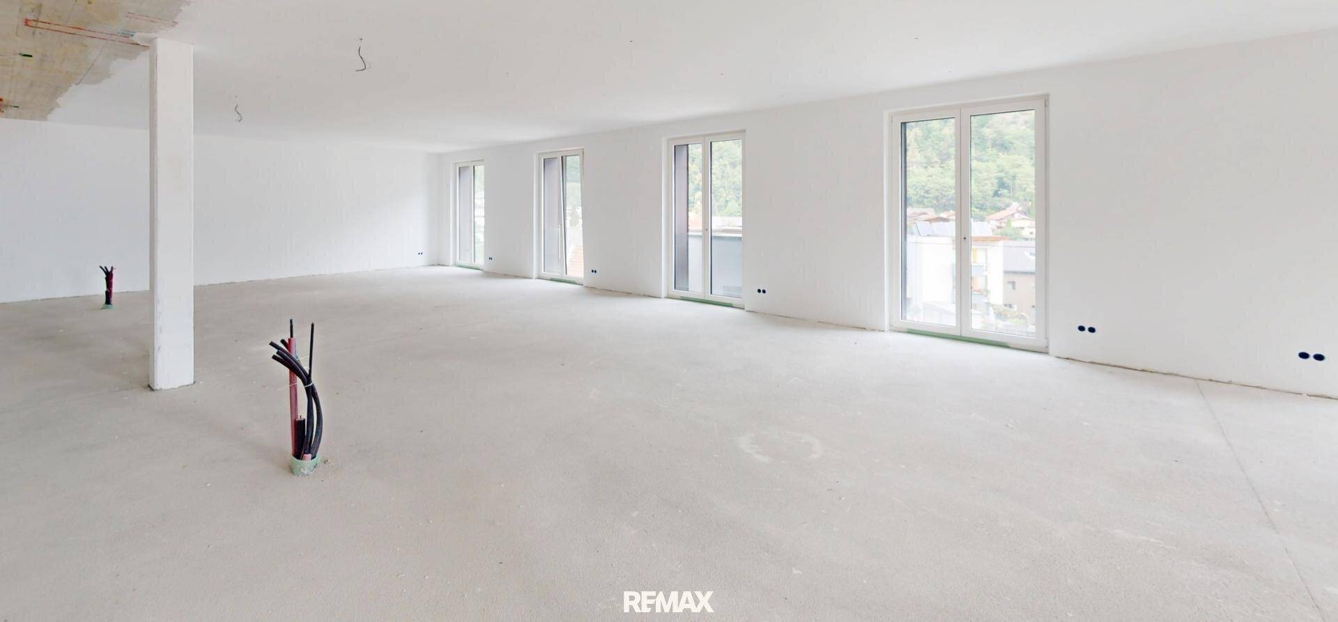Geräumiger, unmöblierter Raum mit Rohinstallationen und großen Fenstern, die viel Tageslicht hereinlassen.