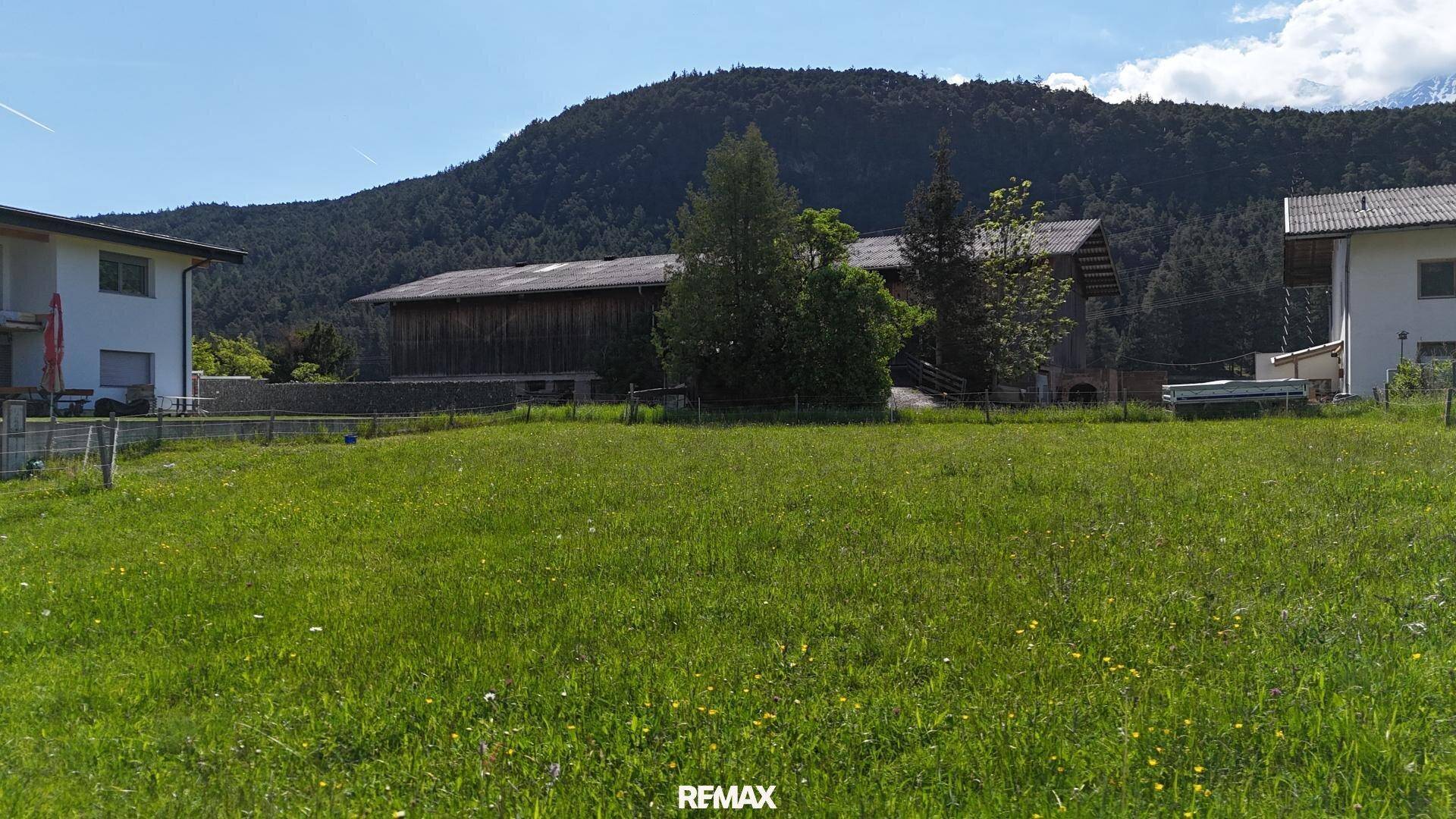 Grüne Wiese mit ländlichen Gebäuden und einem bewaldeten Hügel im Hintergrund unter blauem Himmel.