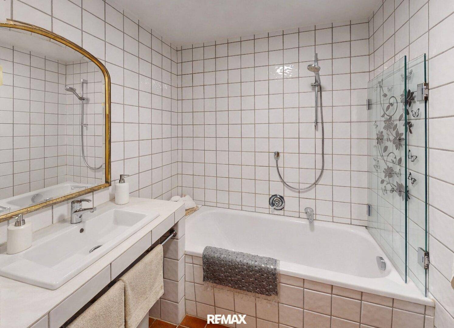 Badezimmer mit weißen Fliesen, Badewanne mit Duschfunktion und einem einzelnen Waschbecken mit goldenem Spiegel.
