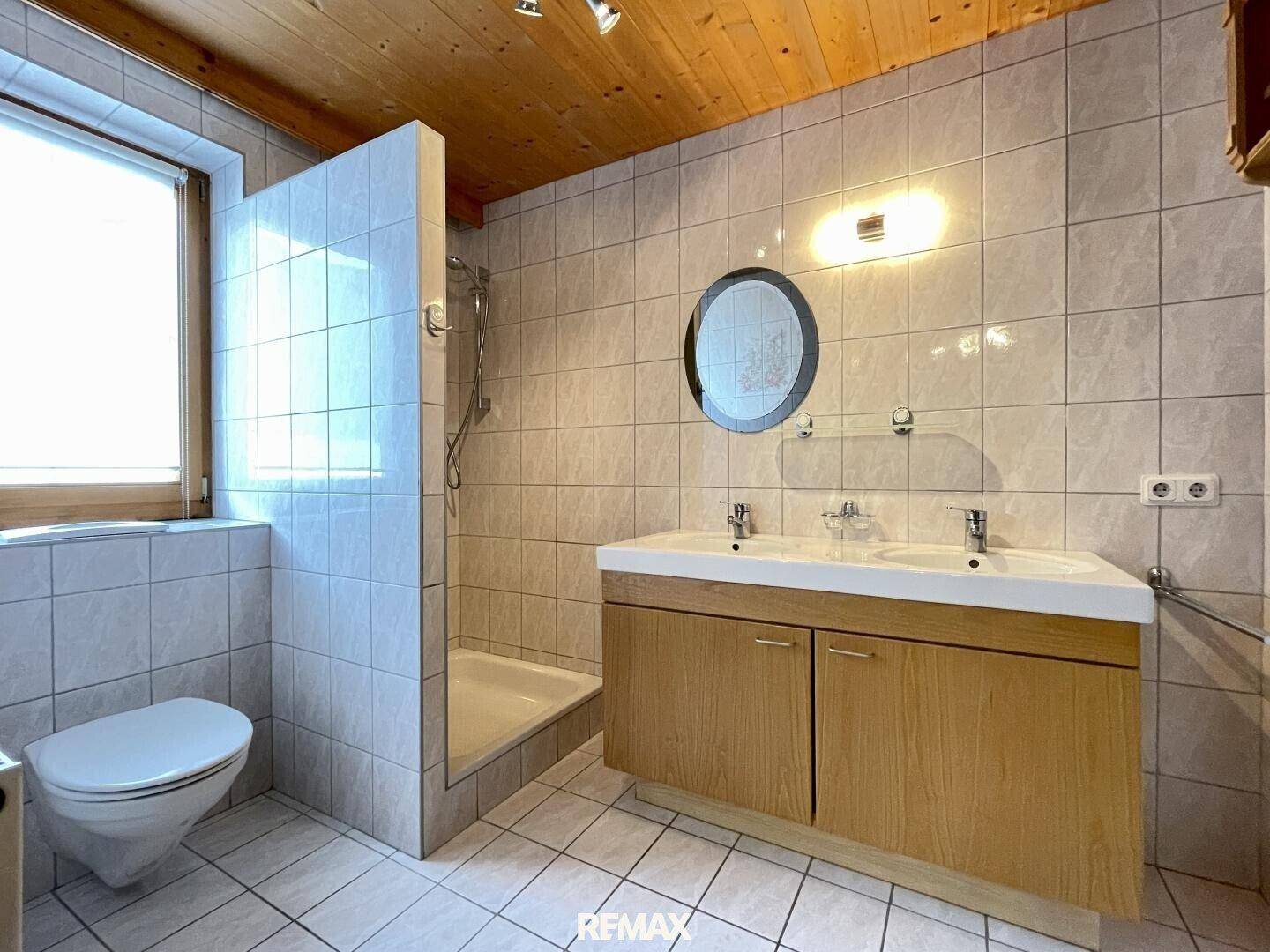 Badezimmer mit Doppelwaschbecken, Dusche und Fenster, gefliest in hellen Tönen.