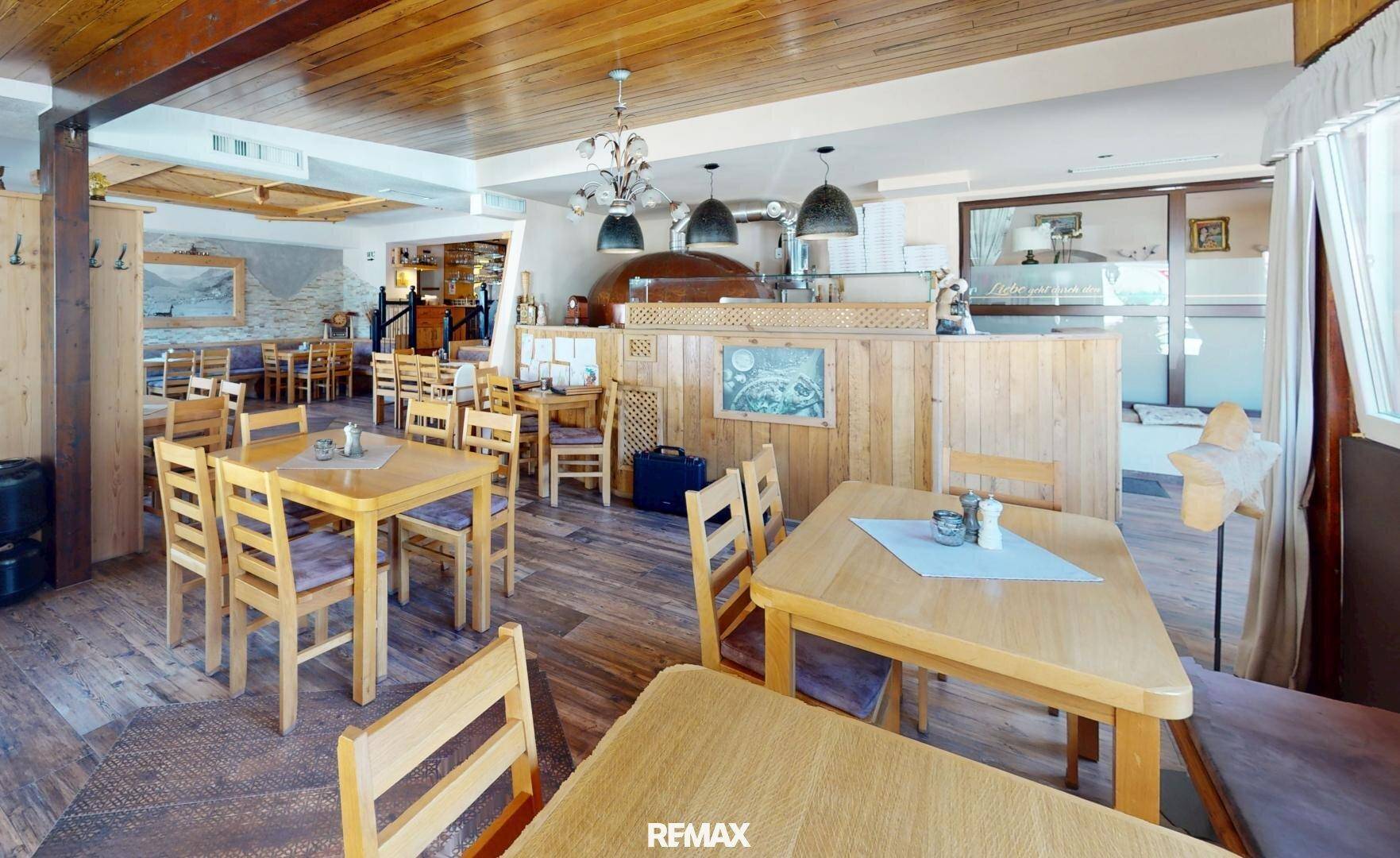 Gemütlicher Speisesaal mit Holztischen und -stühlen, angrenzend an den Bereich mit dem Pizzaofen.