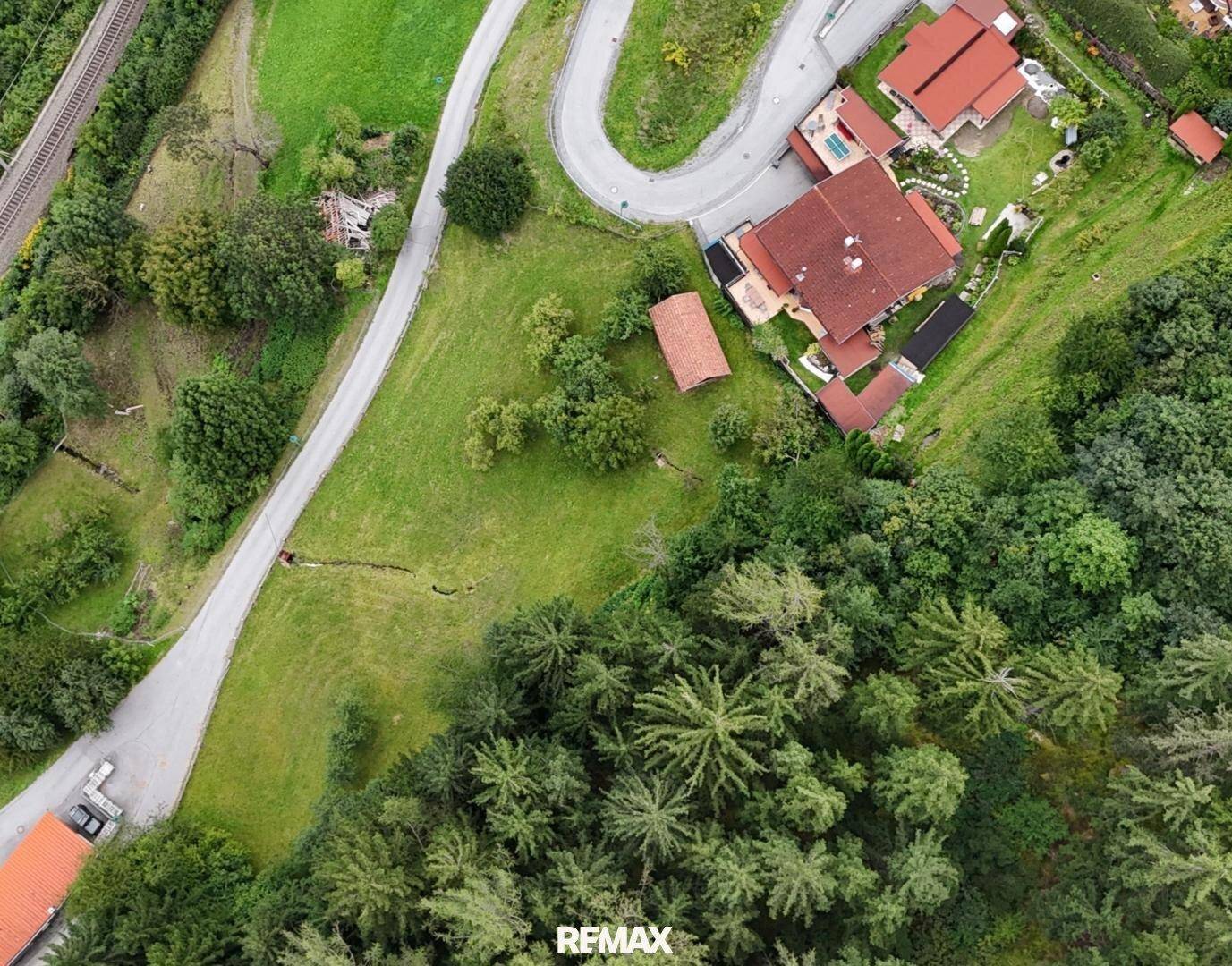 Luftaufnahme einer kurvenreichen Straße, Häusern mit roten Dächern, grünen Feldern und dichtem Wald.