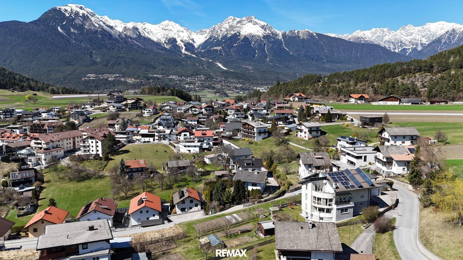 Luftaufnahme eines alpinen Dorfes mit zahlreichen Häusern, umgeben von grünen Wiesen und schneebedeckten Bergen.