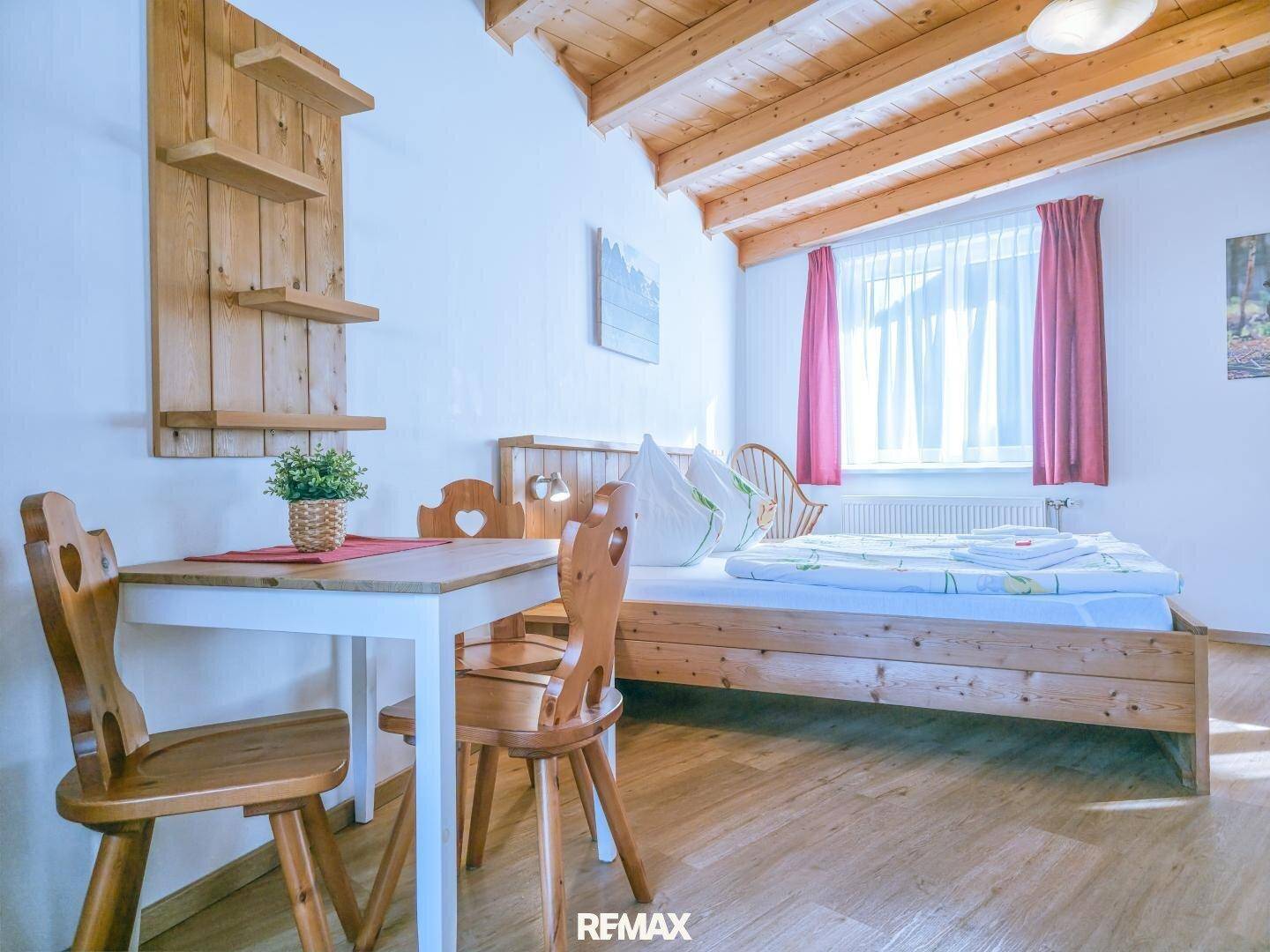 Helles Schlafzimmer mit Holzbett, kleinem Tisch und sichtbaren Holzbalken an der Decke.