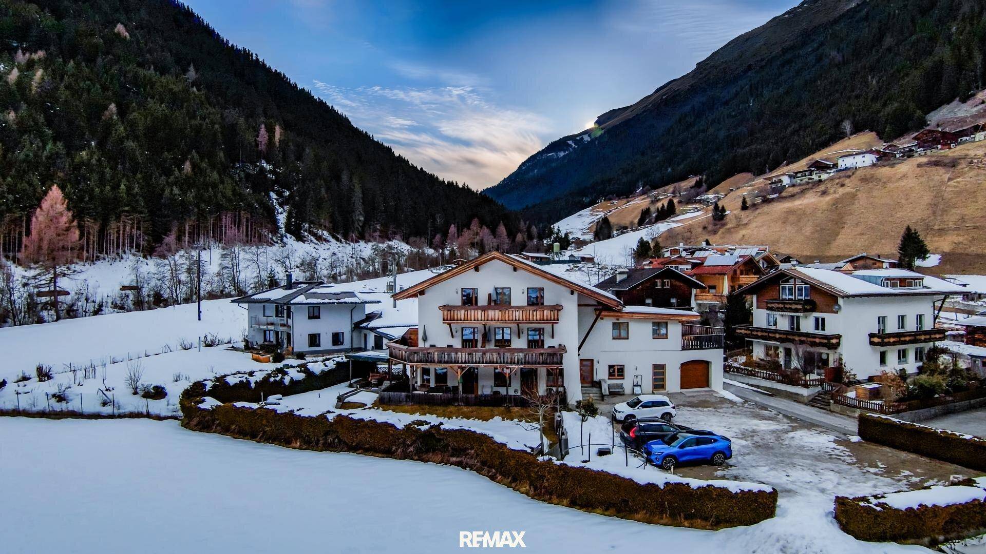 Panoramablick auf ein verschneites Dorf in einem Tal, umgeben von bewaldeten Bergen.