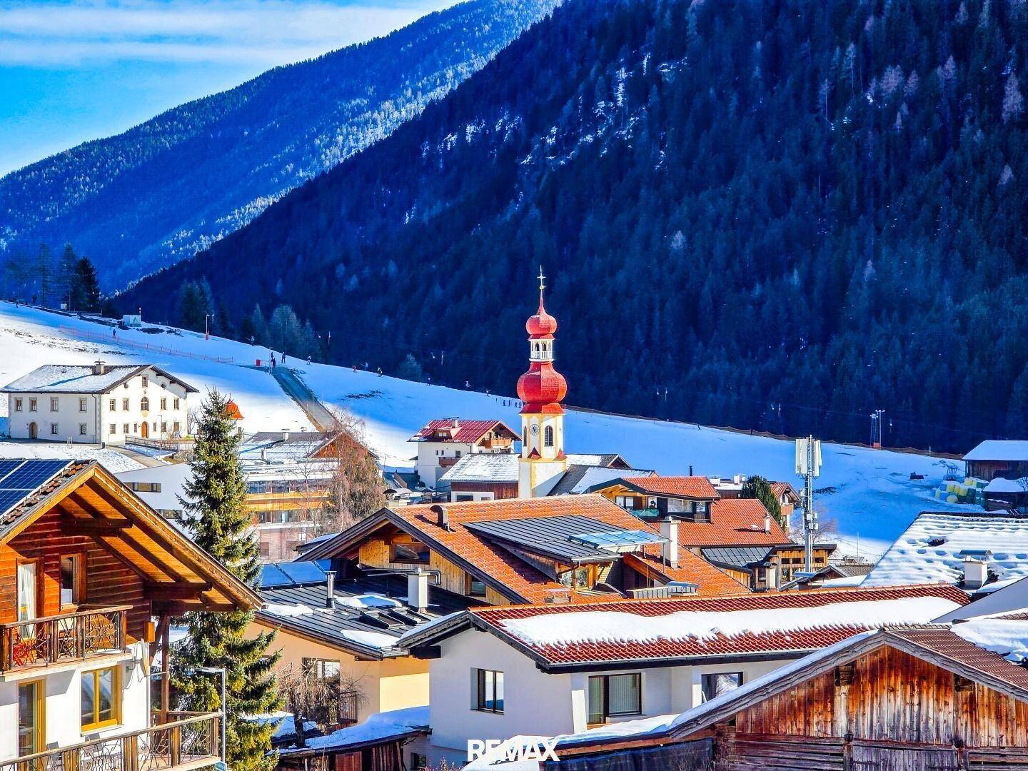 Panoramablick auf ein verschneites Bergdorf mit traditionellen Häusern und einer Kirche unter blauem Himmel.