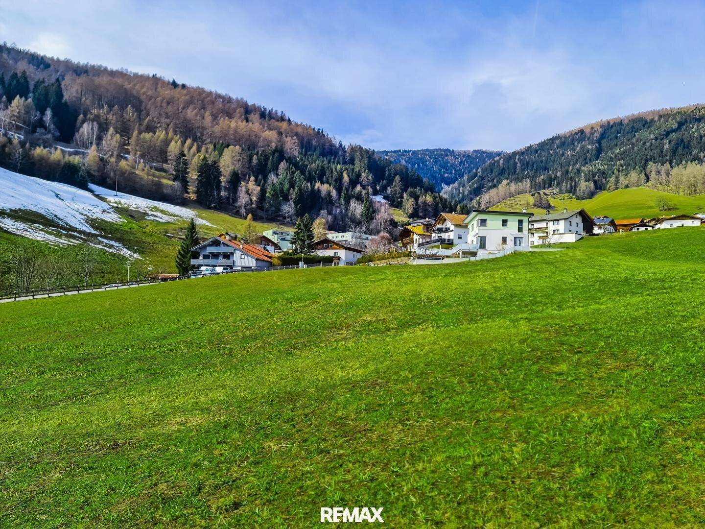 Weitläufige grüne Landschaft mit Häusern im Tal und schneebedeckten Bergen unter blauem Himmel.
