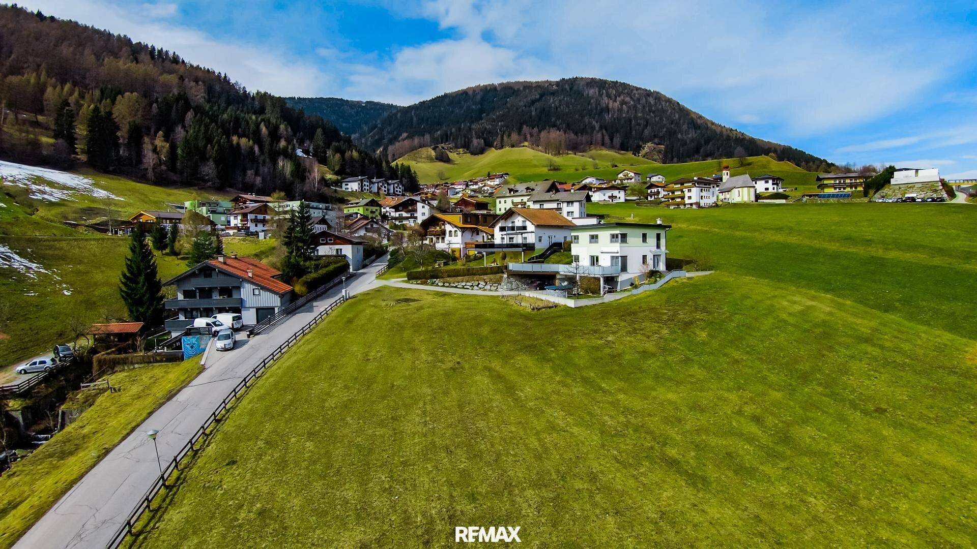 Panoramablick auf ein Dorf in einem grünen Tal, umgeben von bewaldeten Bergen und einem modernen Haus.
