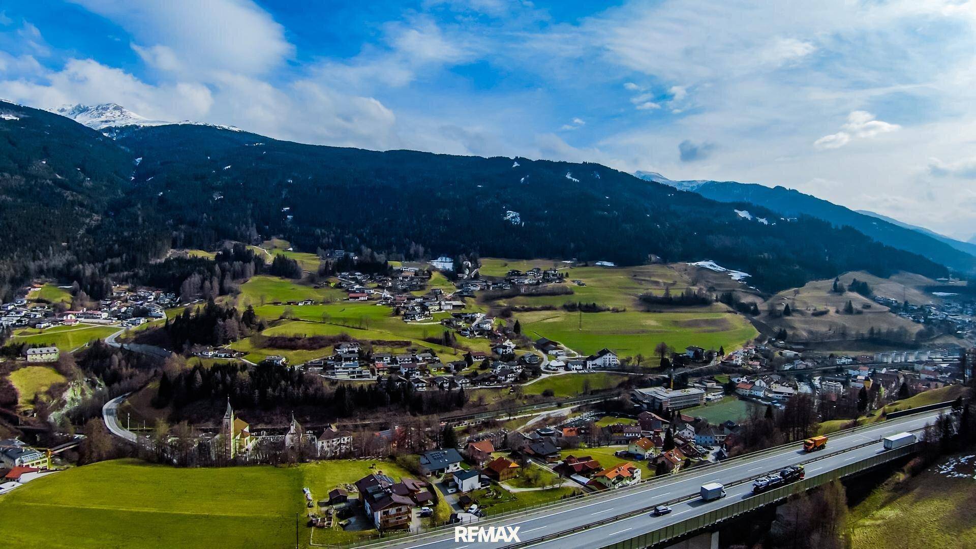 Panoramische Luftaufnahme eines weiten Tals mit Dorf, Autobahnbrücke und majestätischen Bergen im Hintergrund.