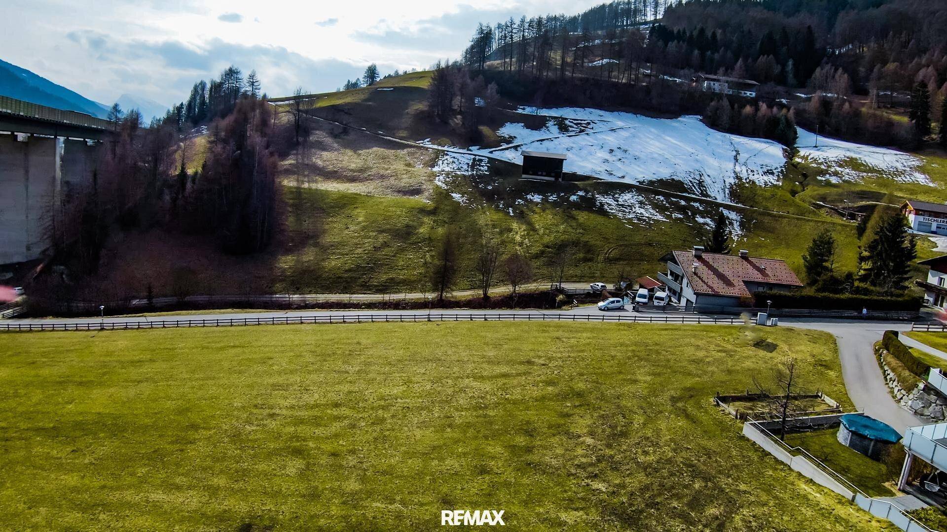 Luftaufnahme eines grünen Feldes mit einem traditionellen Haus und einer Brücke vor schneebedeckten Hügeln.