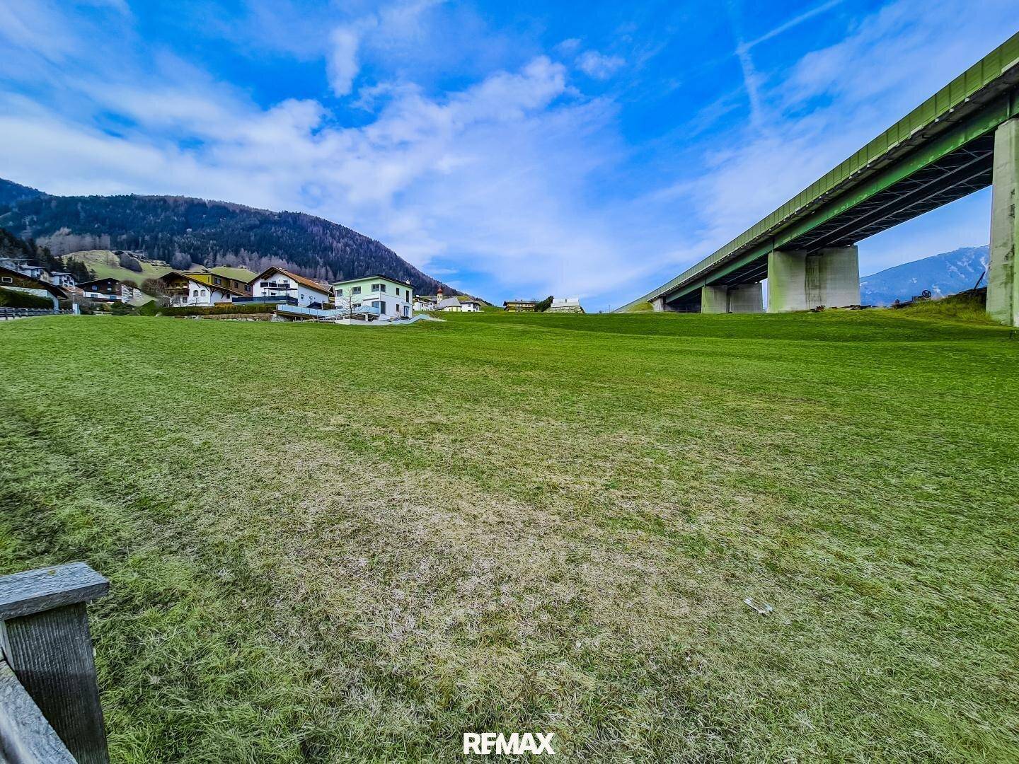 Grüne Wiese mit einer großen Autobahnbrücke und Häusern am Horizont unter einem bewölkten Himmel.