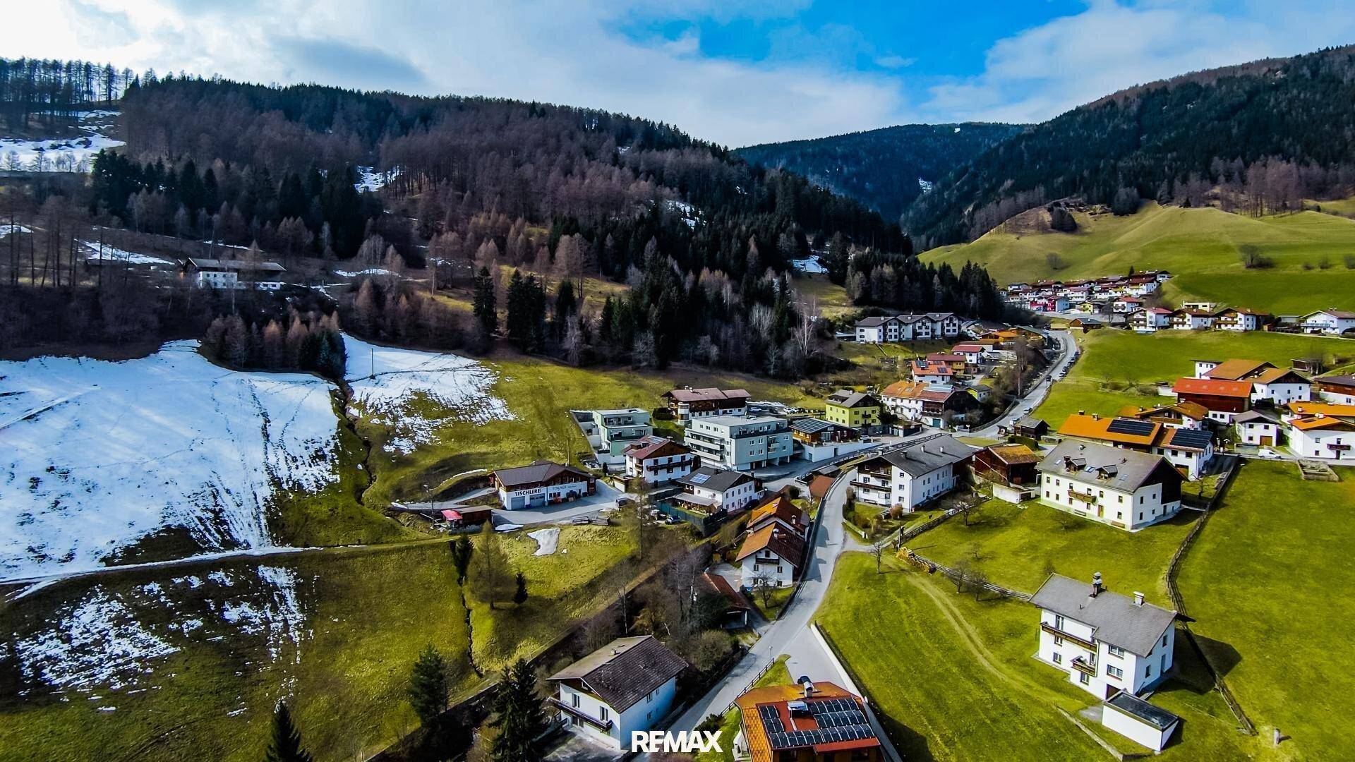 Weitwinkel-Luftaufnahme eines malerischen Dorfes in einem Tal, umgeben von bewaldeten Bergen und Schnee.