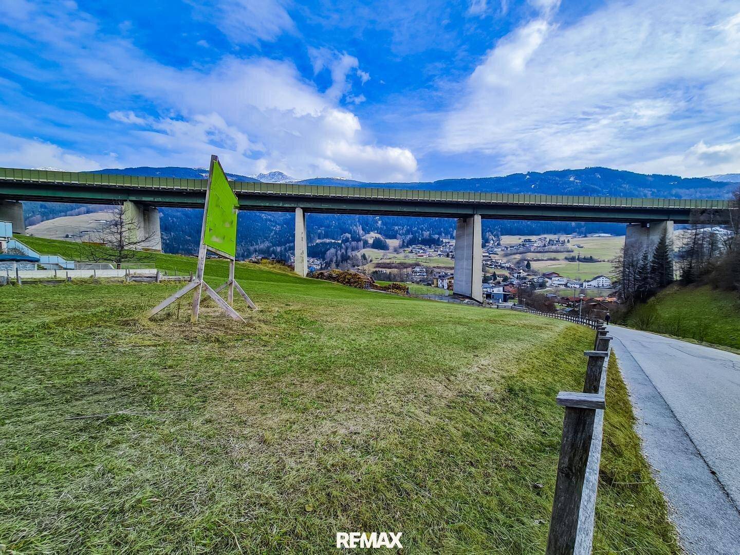Grüne Wiese mit einer imposanten Autobahnbrücke im Hintergrund und einem Hinweisschild am Straßenrand.