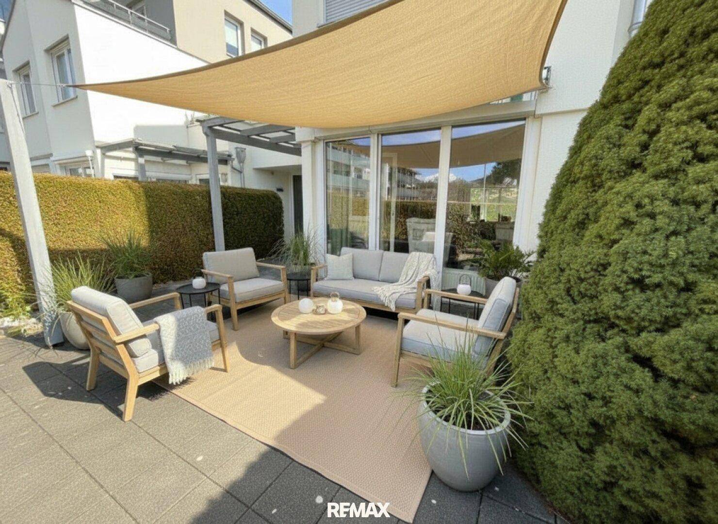 Sonnige Terrasse mit bequemen Gartenmöbeln, Sonnensegel und grüner Hecke im Hintergrund.