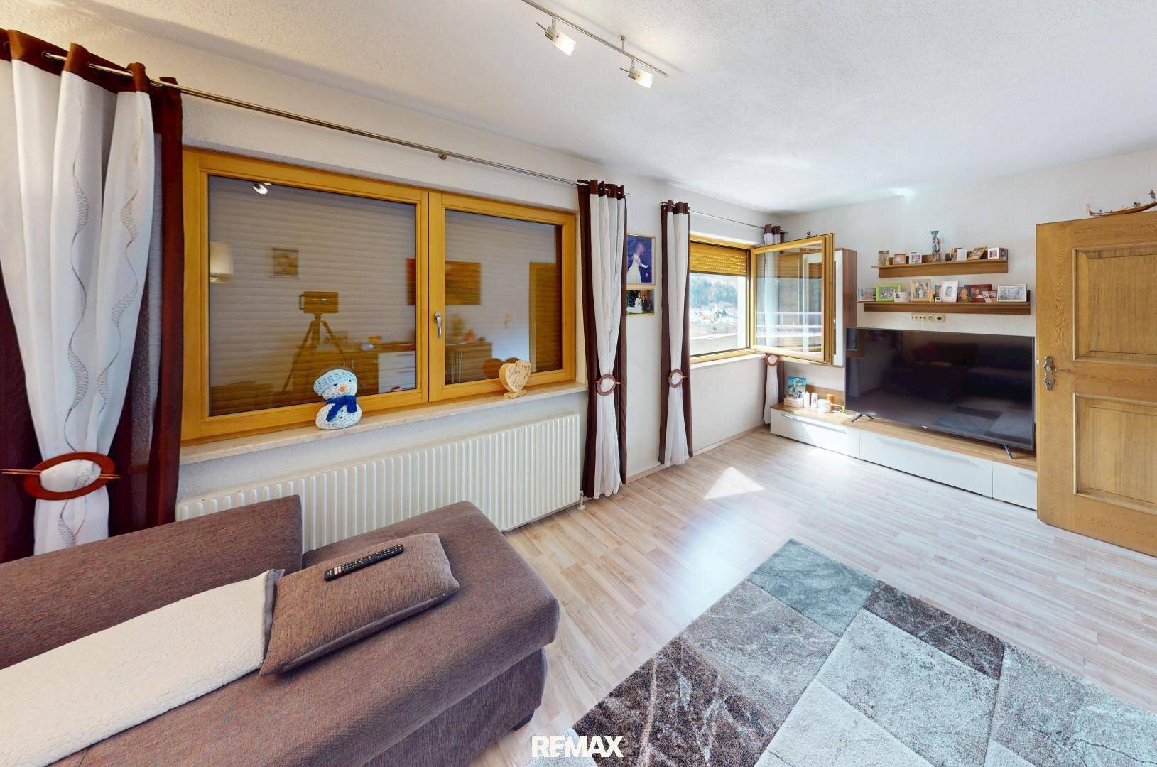 Helles Wohnzimmer mit beigem Sofa, Fernseher und großen Fenstern mit Blick ins Grüne.