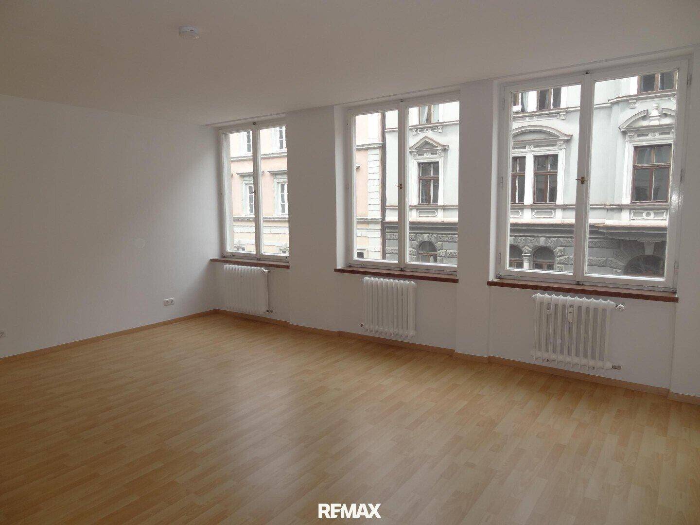 Geräumiger, heller Wohnraum mit drei großen Fenstern, Laminatboden und Blick auf eine historische Fassade.