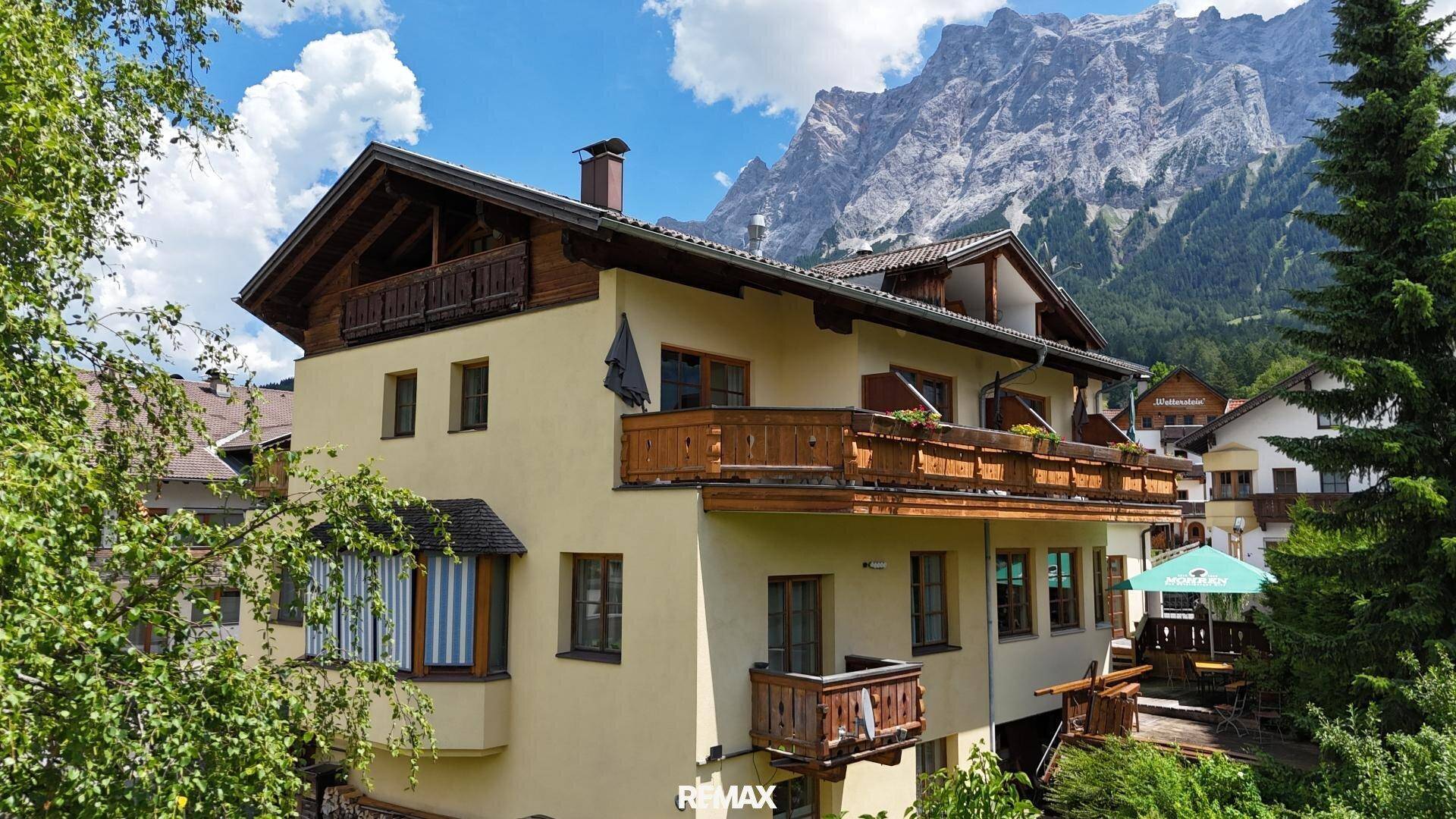 Traditionelles Gebäude mit Holzbalkonen und gelber Fassade, umgeben von Bäumen und Bergen.