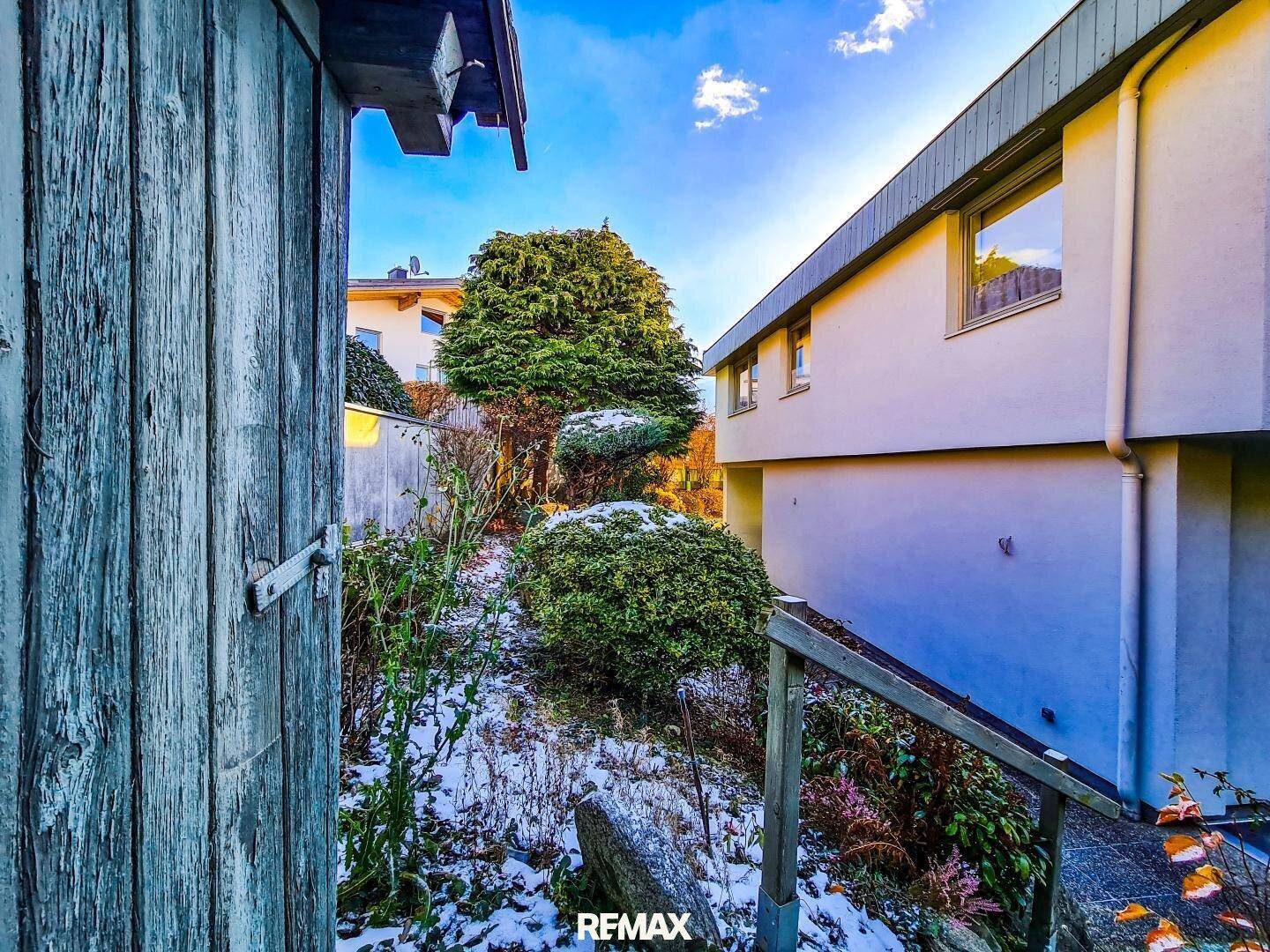 Winterlicher Gartenweg neben dem Haus mit Bäumen und Sträuchern, teilweise mit Schnee bedeckt.