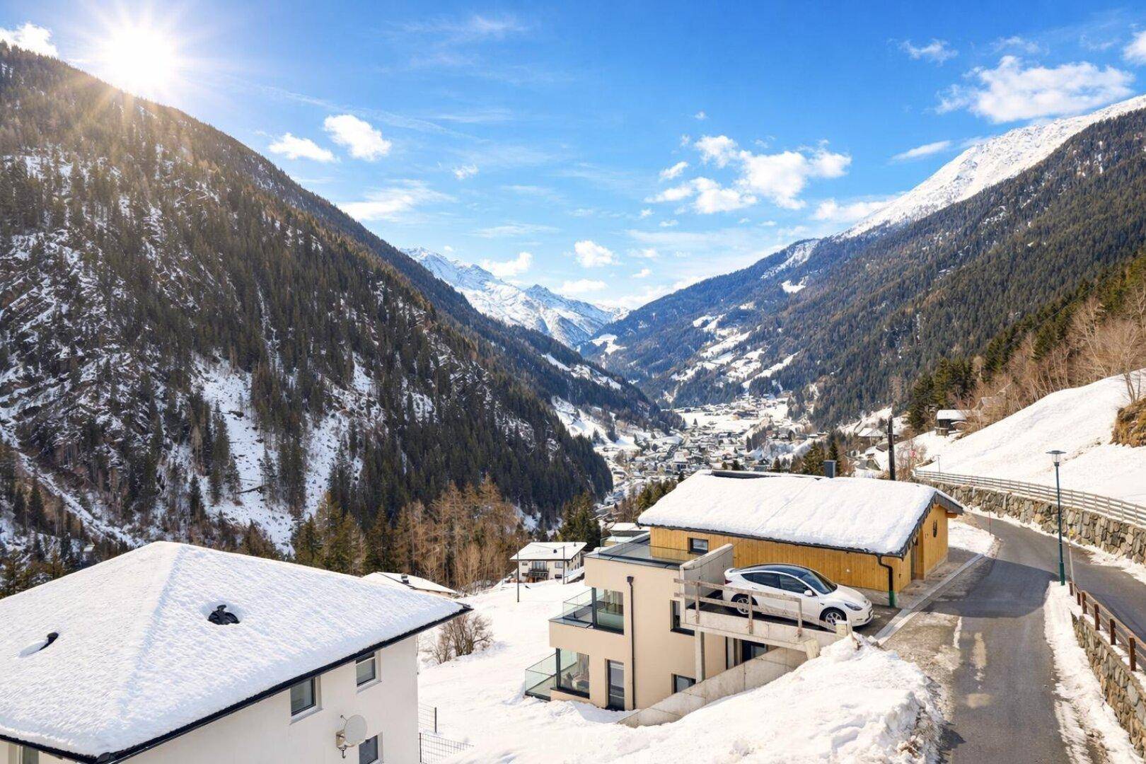 Panoramablick auf verschneite Häuser in einem Tal, umgeben von majestätischen Bergen unter blauem Himmel.