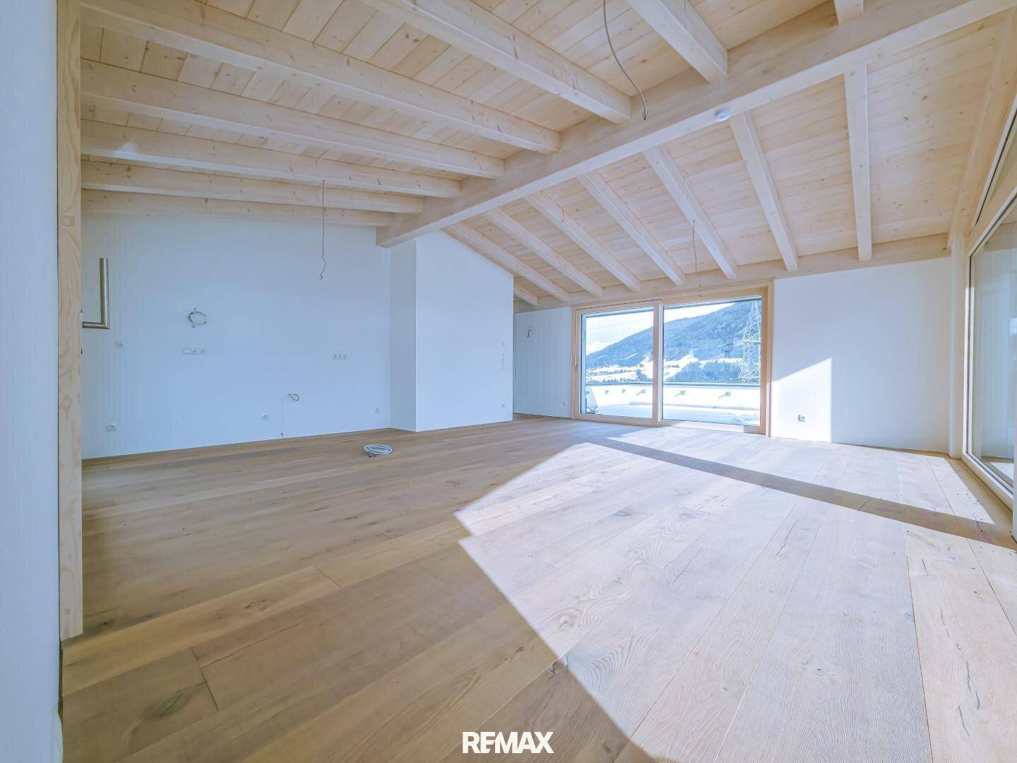 Heller, leerer Raum mit Holzboden und Holzbalkendecke, großen Fenstern und viel Tageslicht.