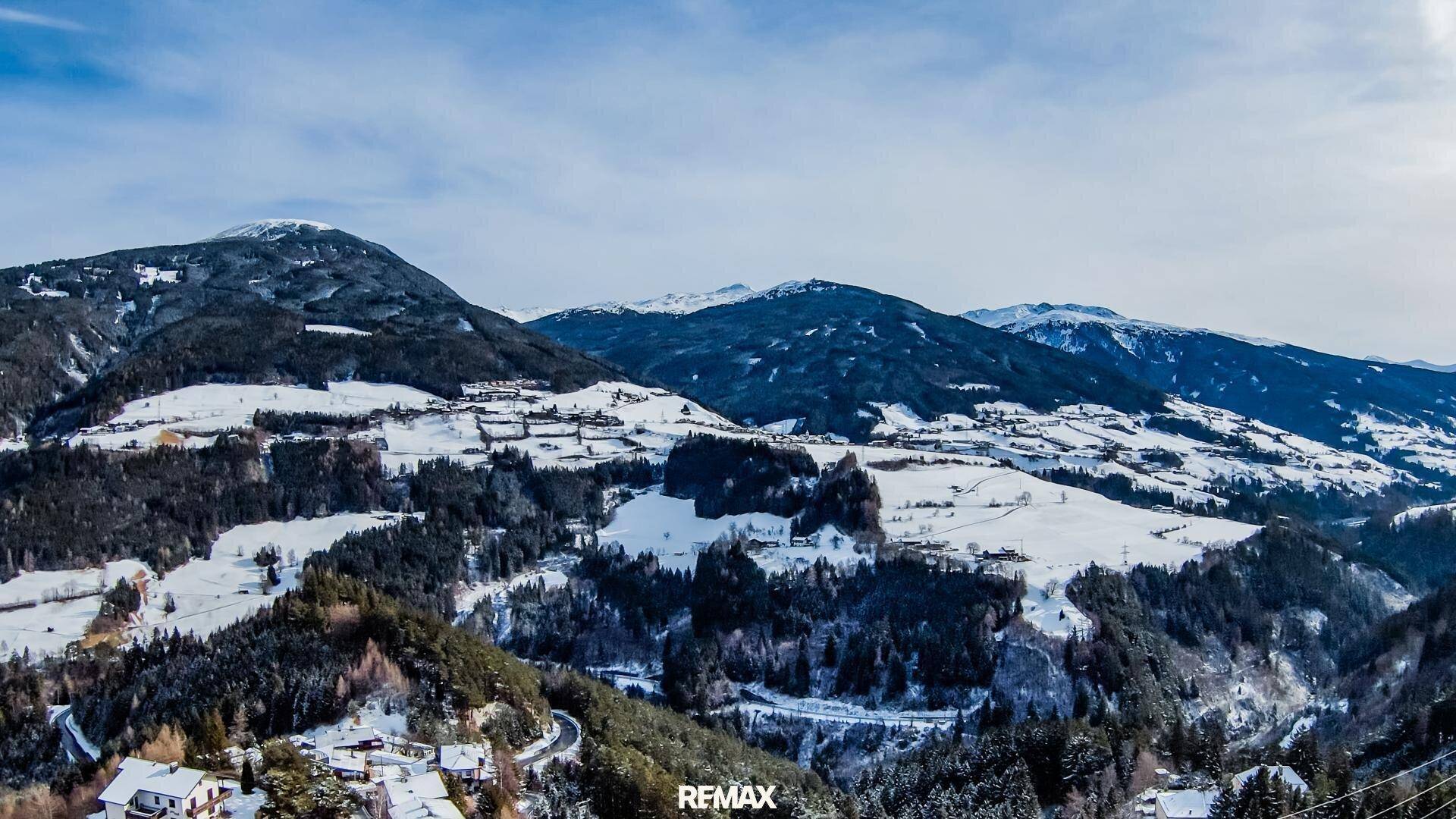 Panoramablick über eine weite, verschneite Berglandschaft mit vereinzelten Häusern und Wäldern.