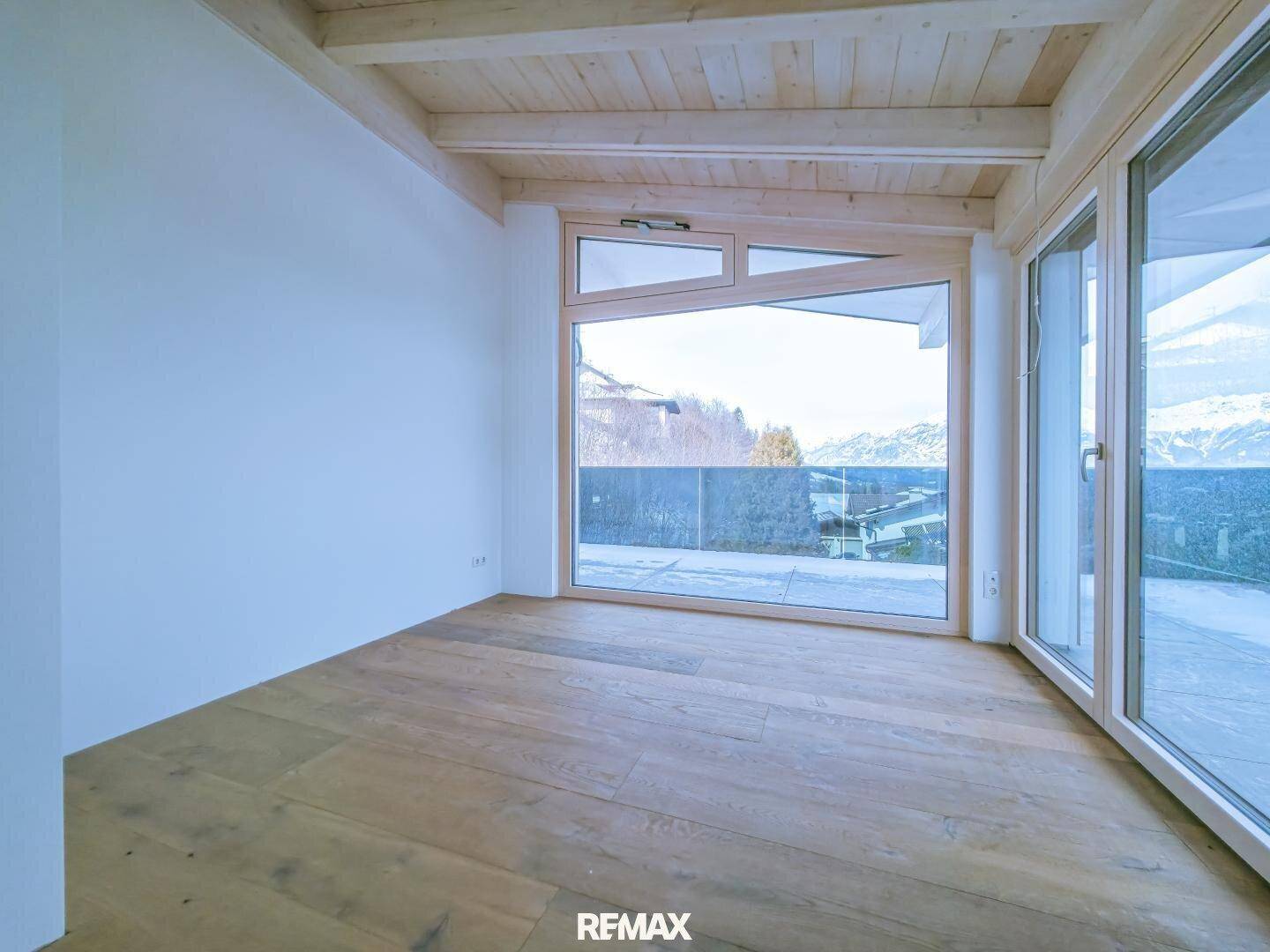 Heller Raum mit Holzboden und Holzbalkendecke, großem Fenster und Blick auf die umliegende Landschaft.