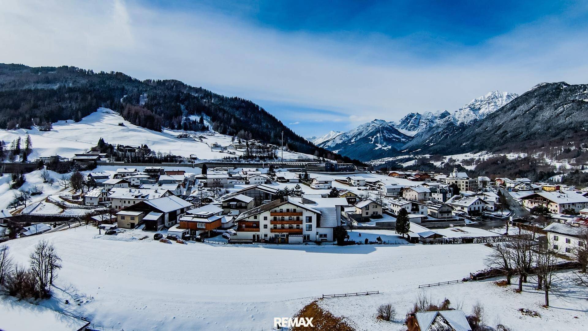 Weitläufiger Blick auf ein verschneites Dorf in einem Tal, umgeben von majestätischen Bergen.