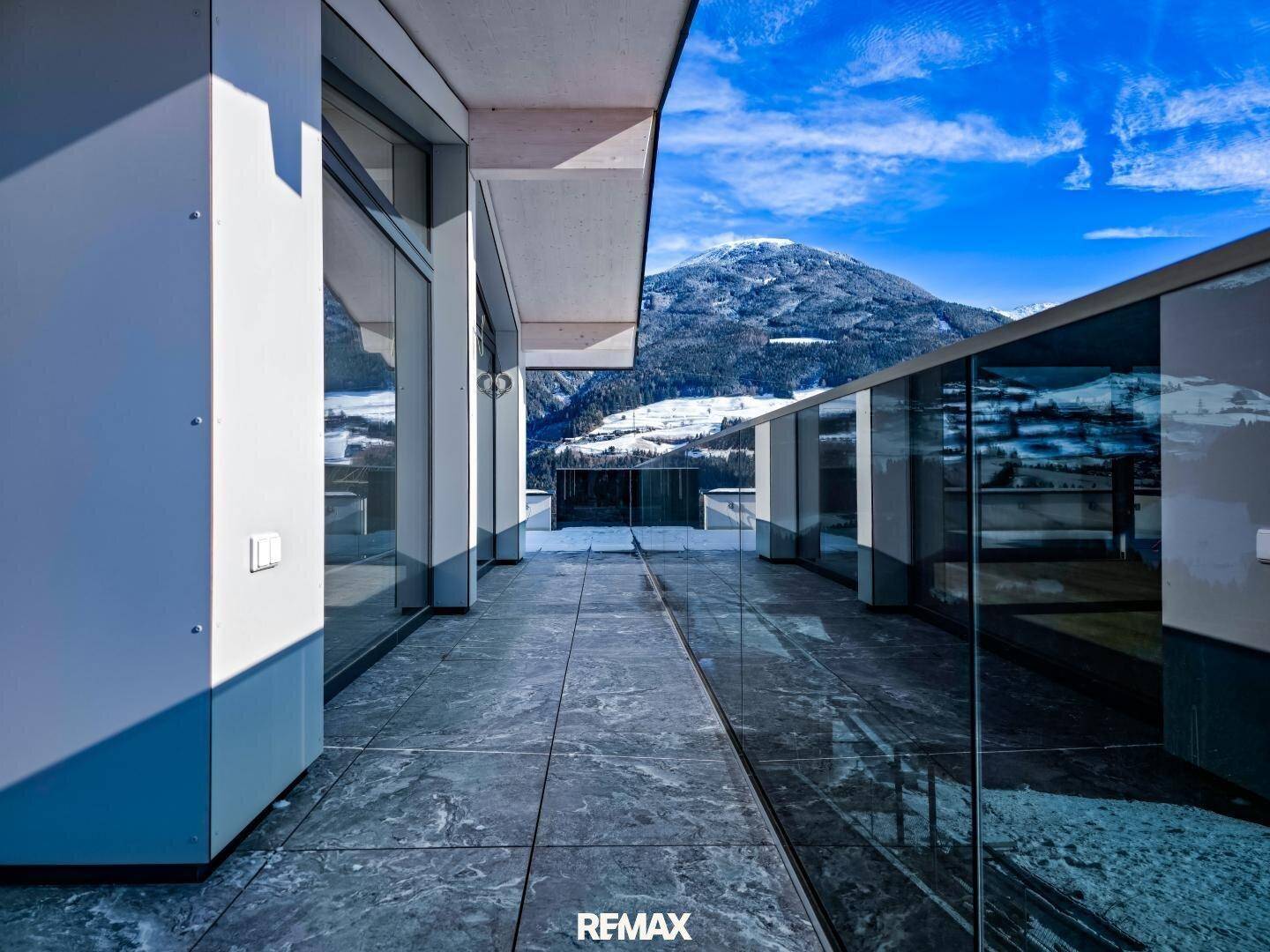 Langer Balkon mit Glasgeländer und Fliesenboden, bietet einen weiten Blick auf die verschneite Bergwelt.