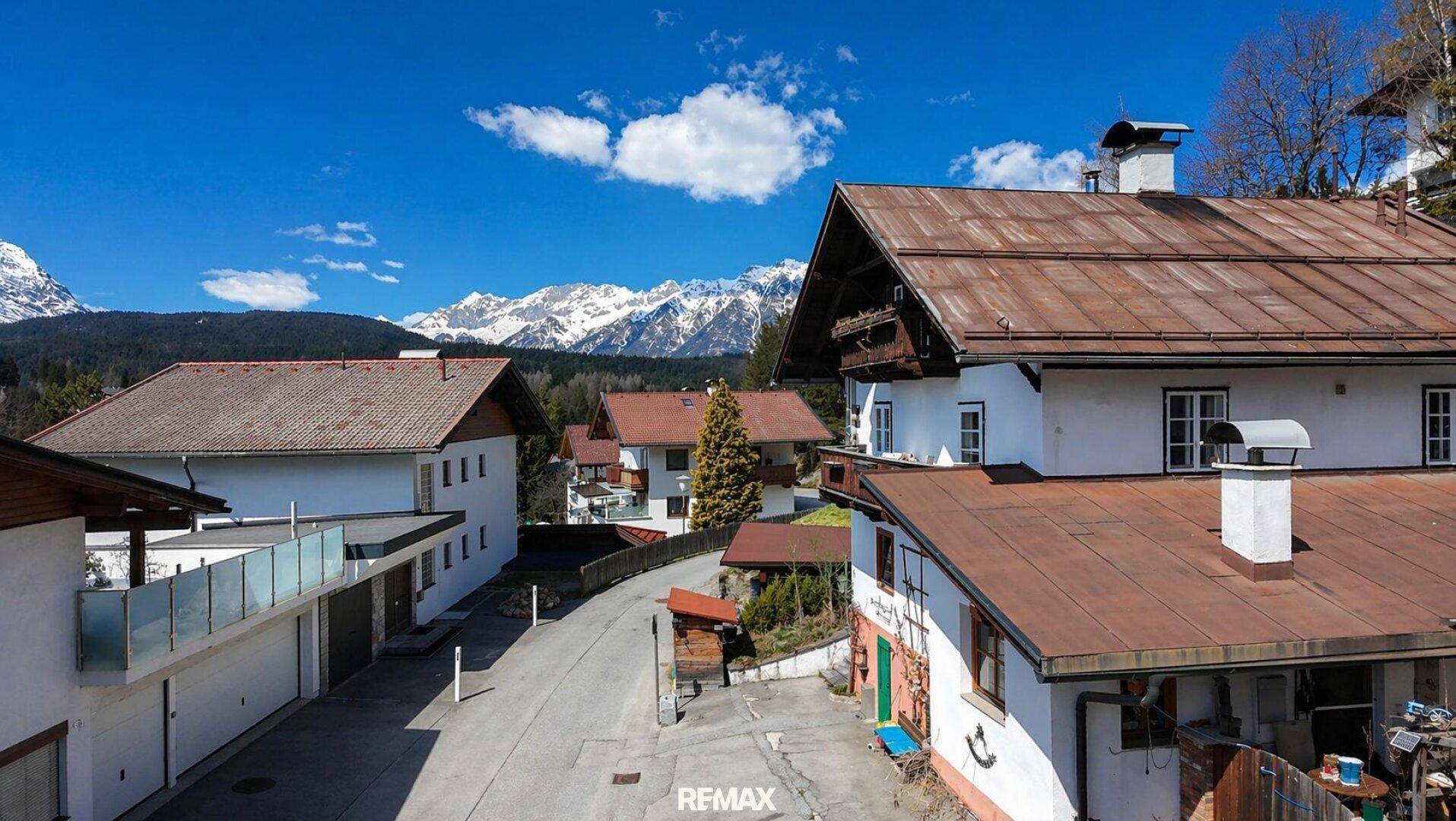 Blick auf eine ruhige Dorfstraße mit traditionellen Häusern und schneebedeckten Bergen im Hintergrund.