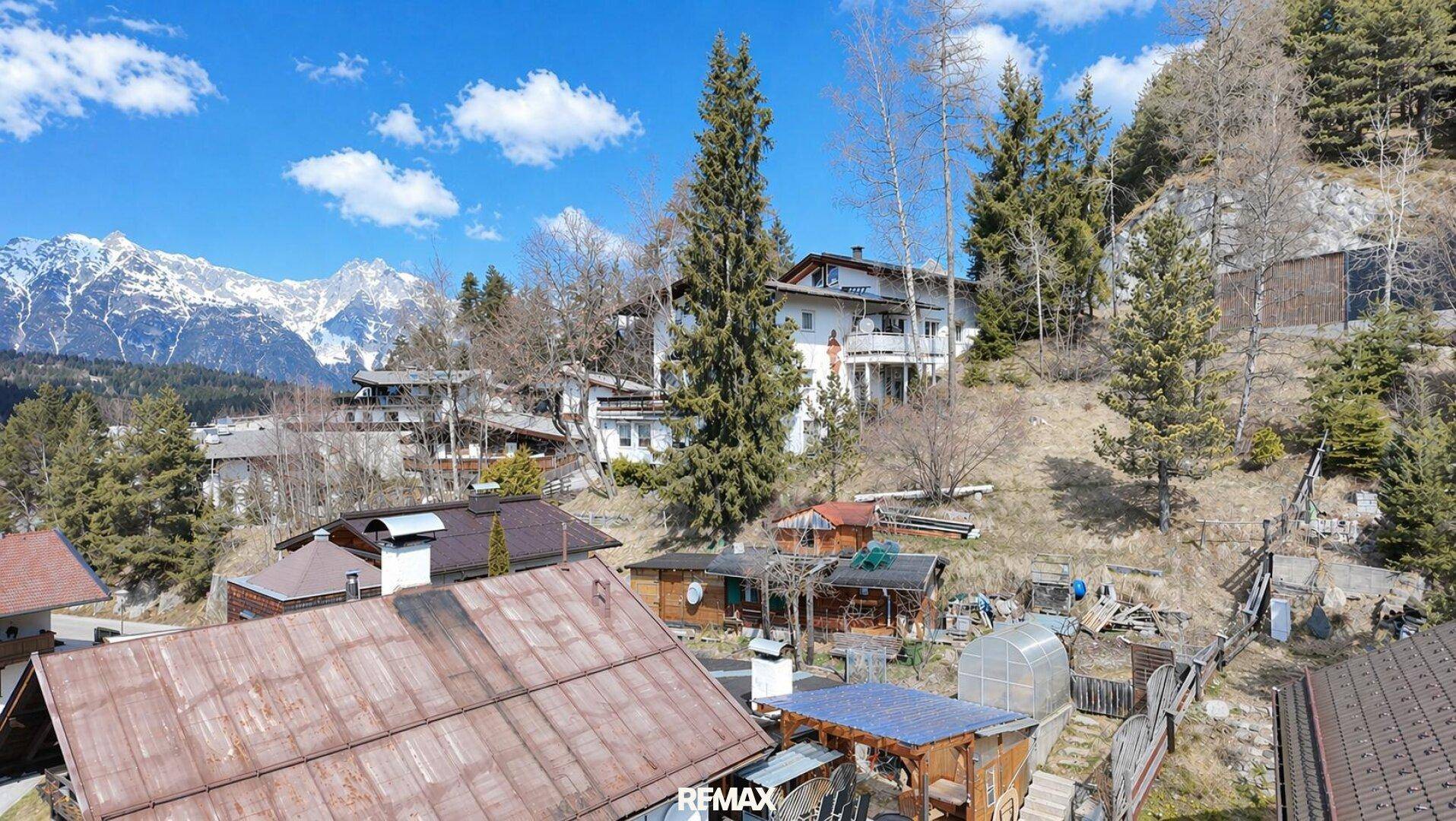 Häuser auf einem sonnigen Hang mit Bäumen und einem weiten Blick auf die Berglandschaft.