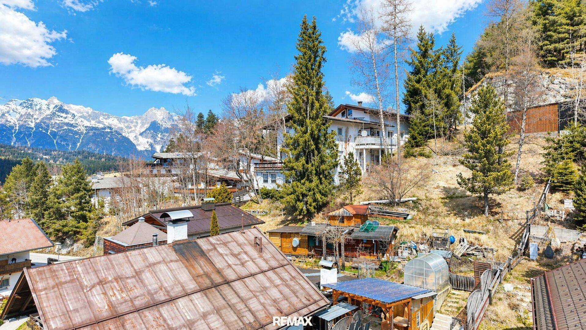 Häuser auf einem sonnigen Hang mit üppiger Vegetation und majestätischen Bergen in der Ferne.