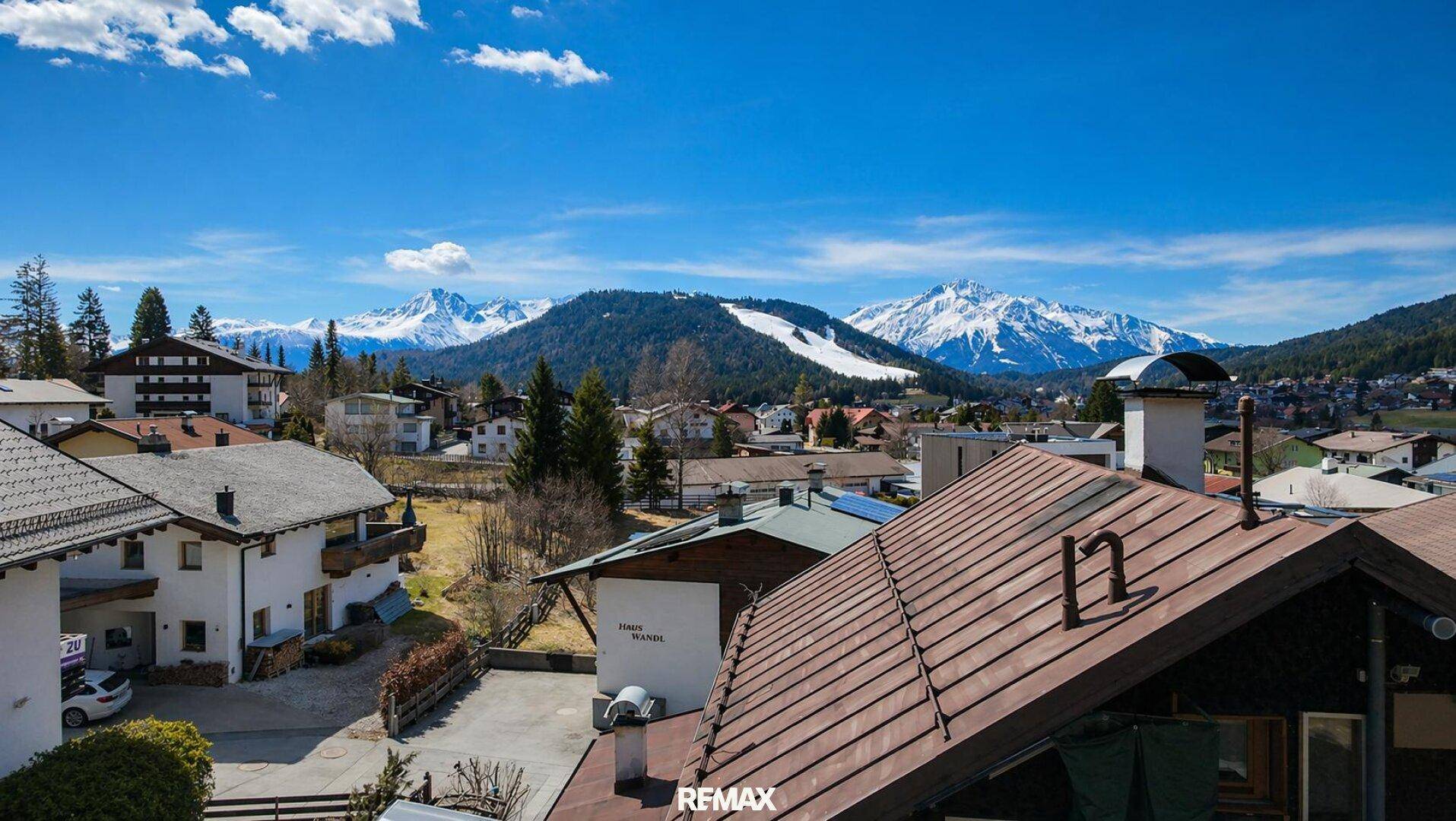 Panoramablick über ein alpines Dorf mit schneebedeckten Bergen und einer Skipiste unter blauem Himmel.