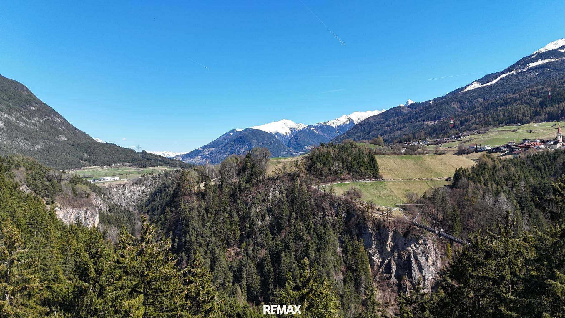 Weitläufige Landschaftsaufnahme mit tiefen Tälern, dichten Wäldern und schneebedeckten Berggipfeln unter blauem Himmel.